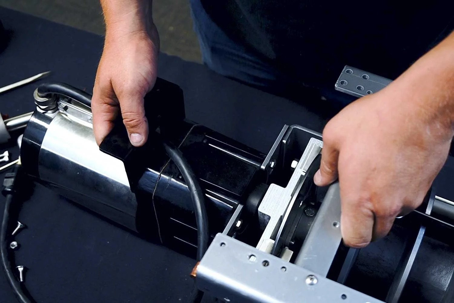 Hands assembling or repairing a mechanical or electronic device with metal and black components on a black surface.
