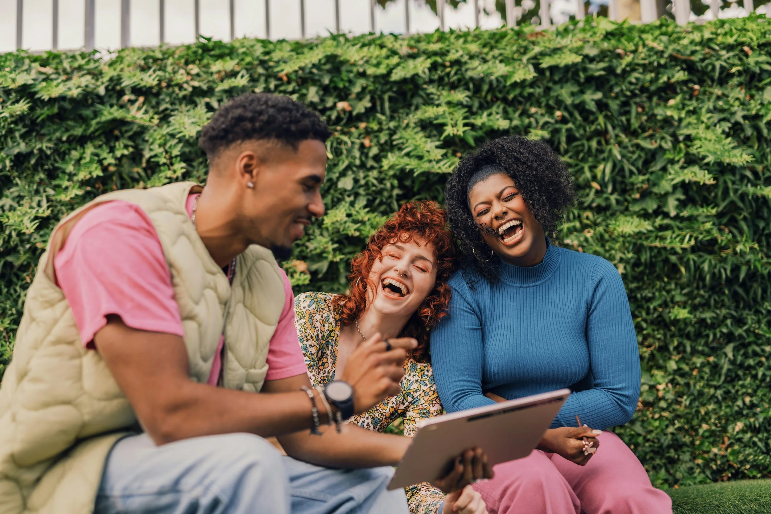 Four friends sitting outdoors on a bench, laughing and looking at a tablet, with green bushes and a metal fence in the background.
