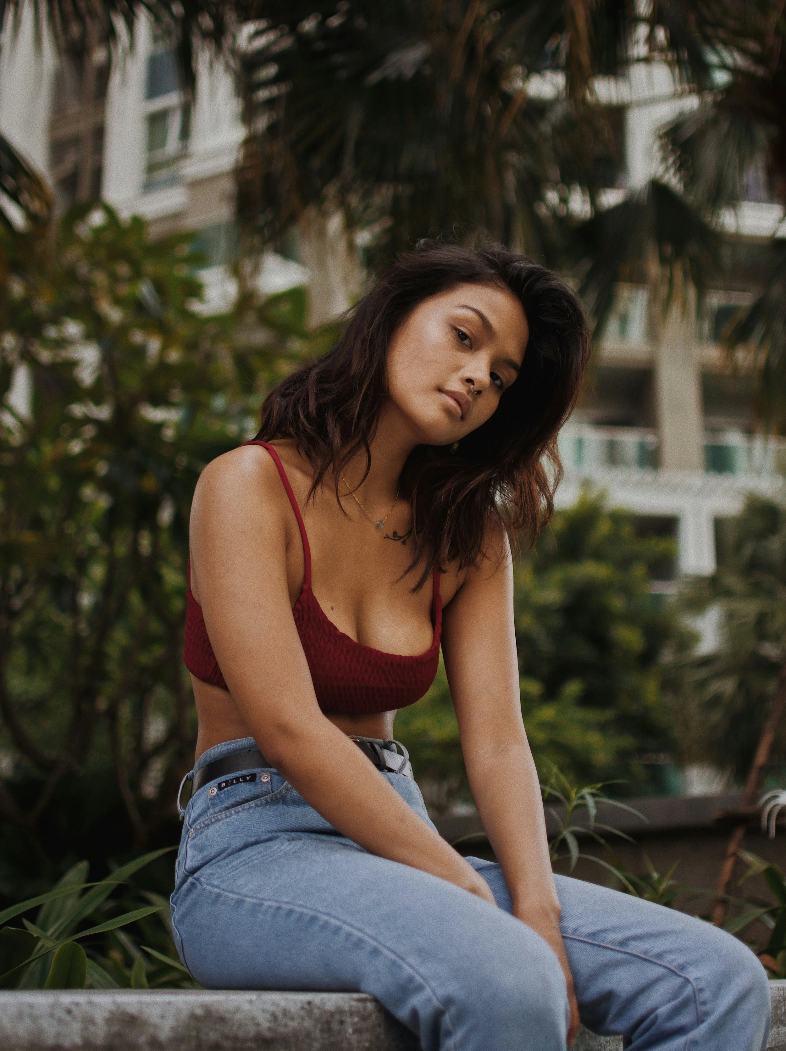 A young woman with dark wavy hair, wearing a red crop top and blue jeans, sitting outdoors on a concrete ledge with greenery and a building with balconies in the background.
