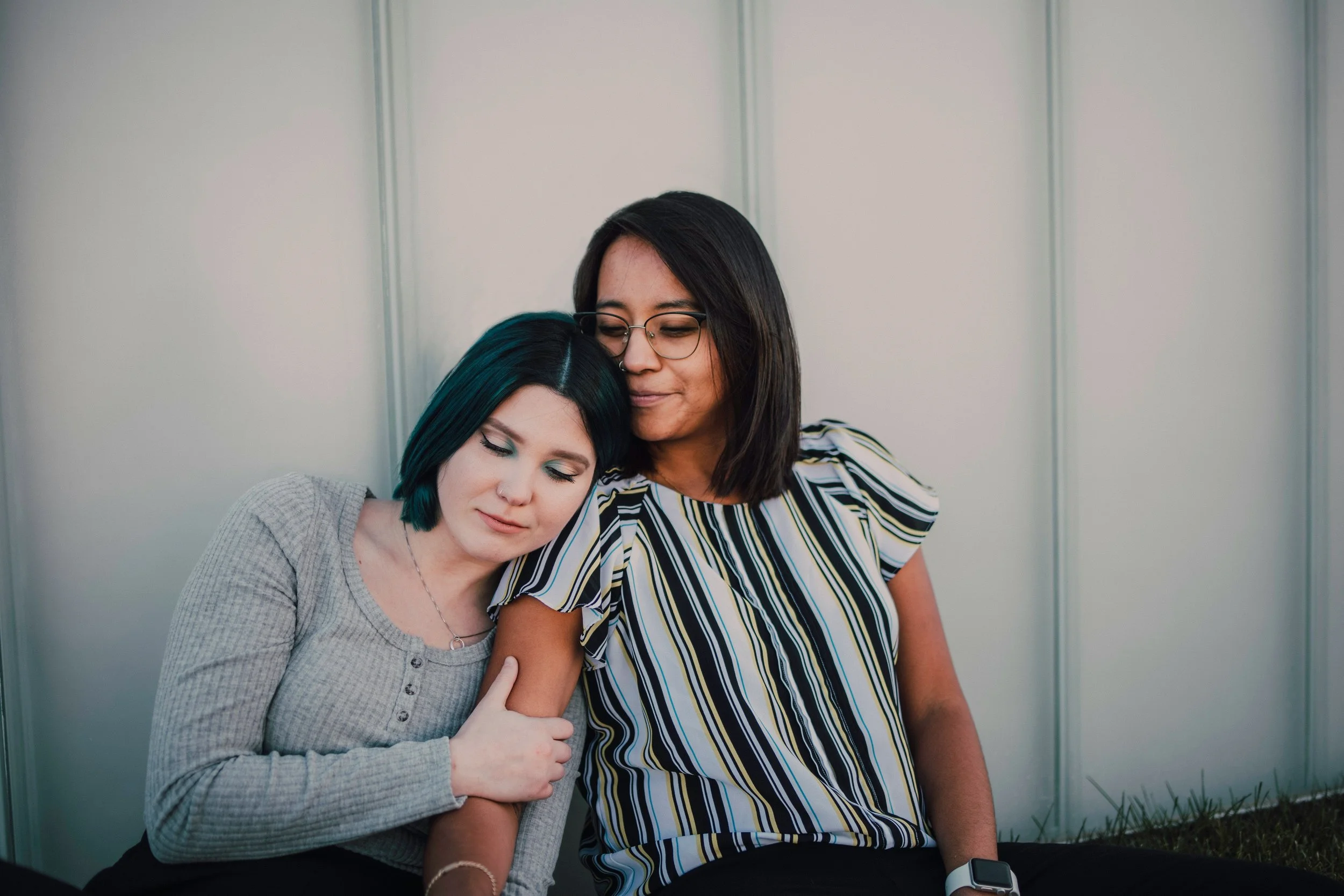 Two women sitting close together against a gray wall; one with long dark hair, glasses and striped shirt, embracing and resting her head on the shoulder of the other woman with short teal hair and light gray cardigan, with eyes closed.