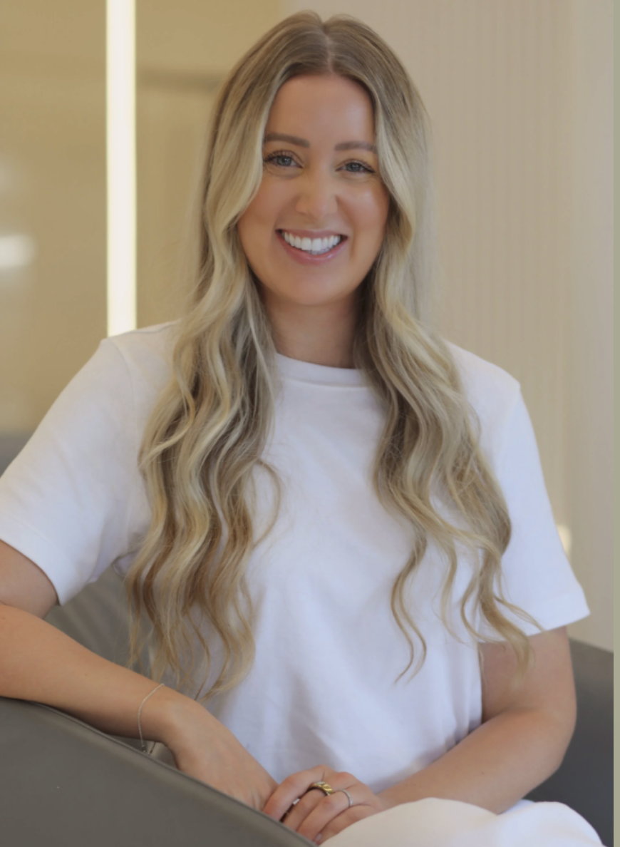 A smiling woman in trauma therapy with long blonde hair wearing a white t-shirt, sitting on a gray chair in a well-lit indoor space.
