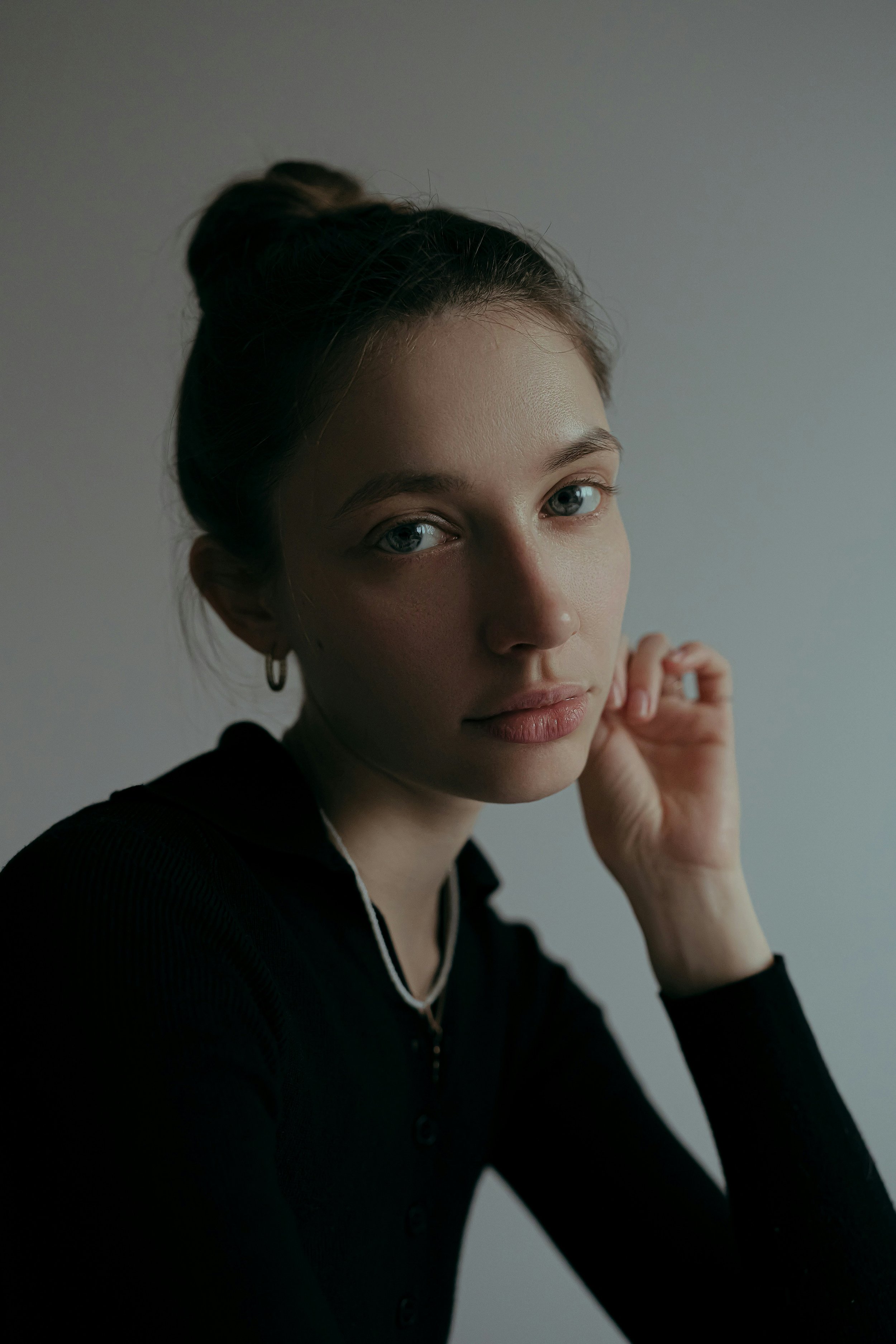 A woman with blue eyes and brown hair styled in a bun, wearing a black top with white piping, earrings, and a necklace, is looking directly at the camera, with her hand gently touching her face after going to therapy for chronic illness in california