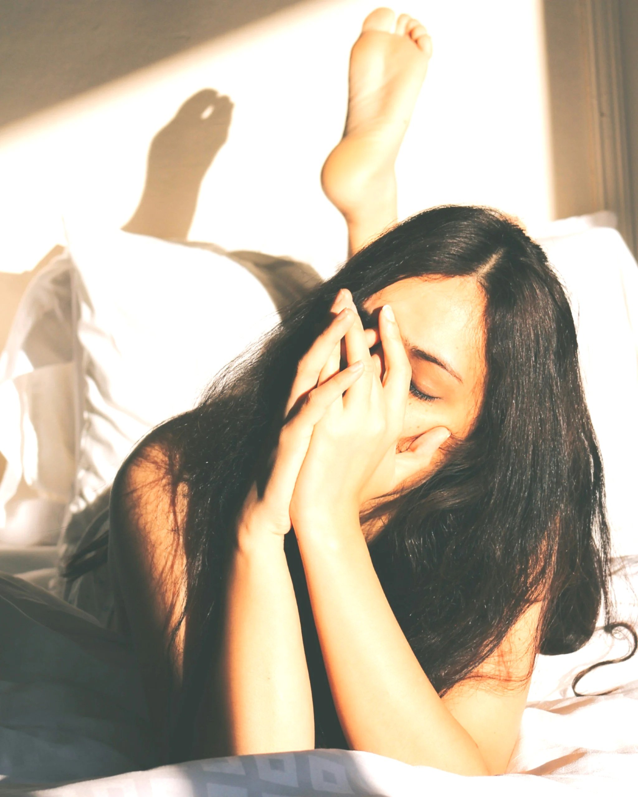 A woman with long dark hair covering her face, lying on a bed with sunlight casting shadows and a hand near her face.