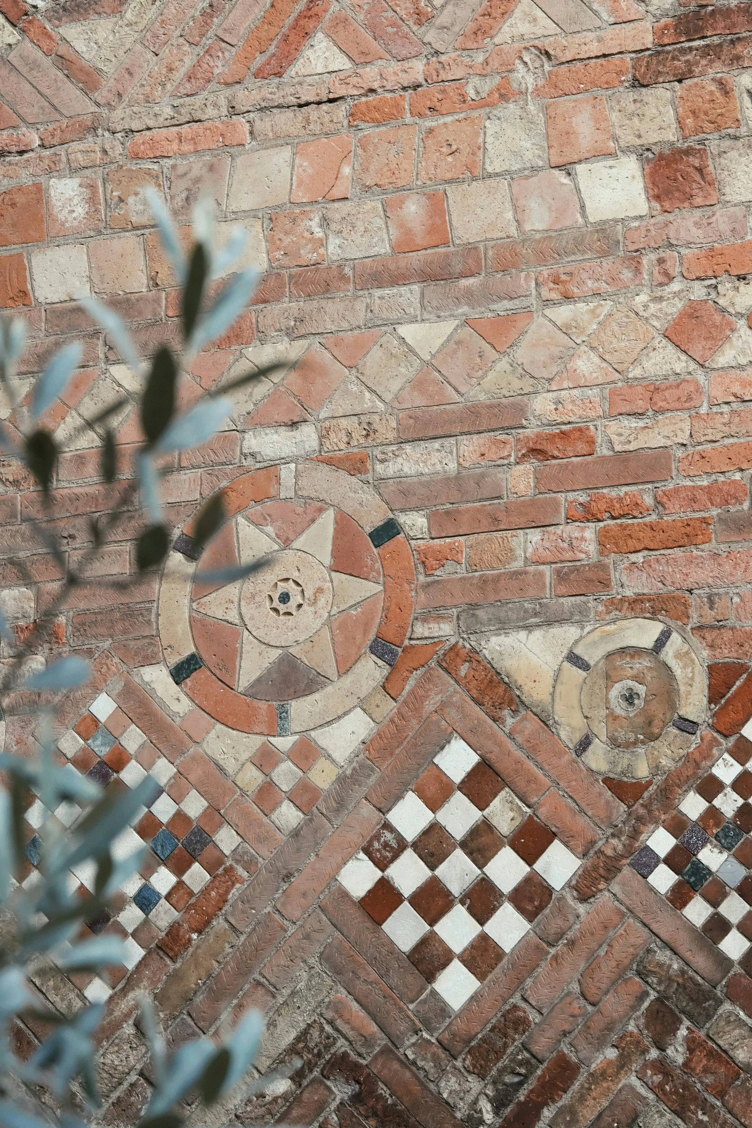Close-up of an intricate brick and tile mosaic on a wall and floor, with some greenery in the foreground.