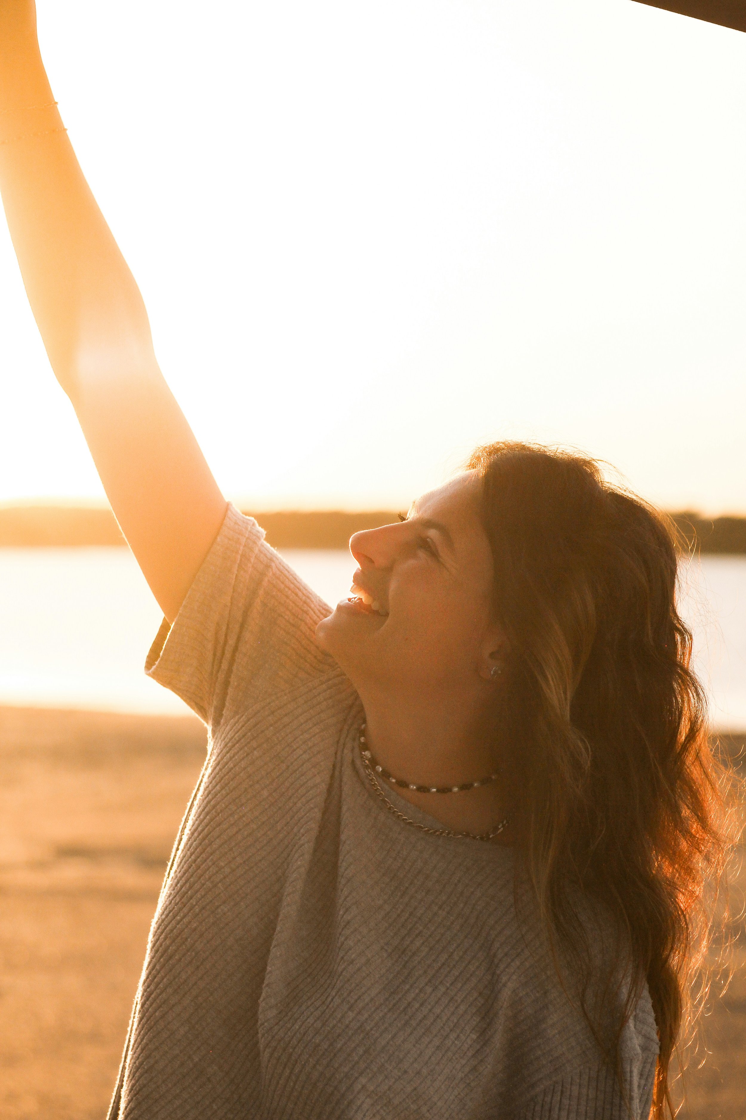 A young woman with long hair smiling and raising her arm in a relaxed outdoor setting at sunset with a body of water in the background.
