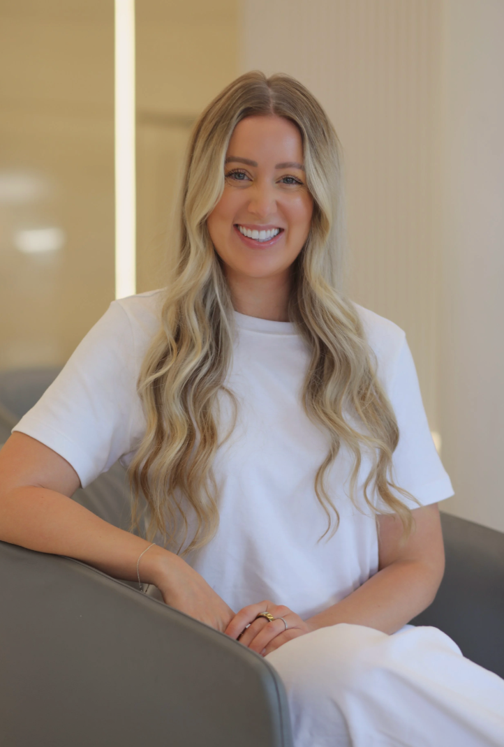 A woman with long blonde wavy hair smiling and sitting on a gray chair in a light-colored room.