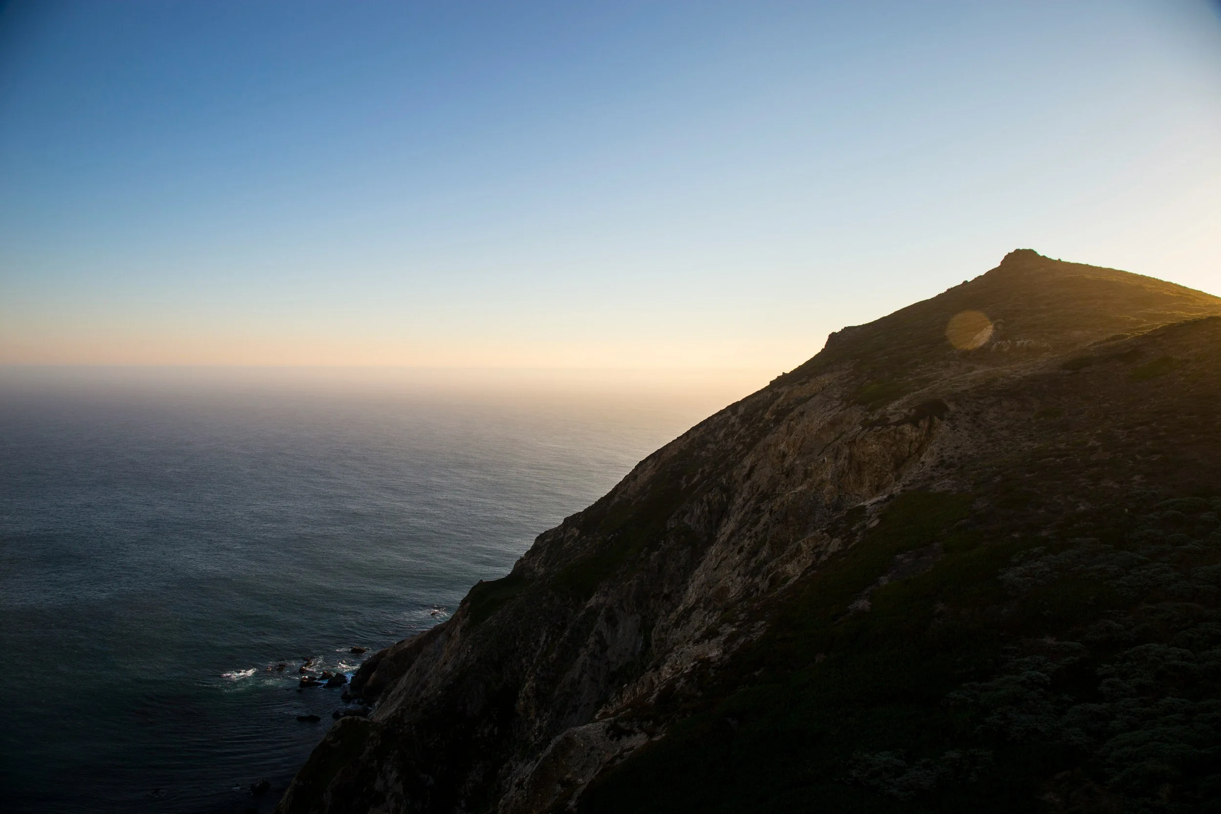 A coastal scene with a rugged hill or mountain slope descending to the ocean, illuminated by soft sunlight at sunset or sunrise.