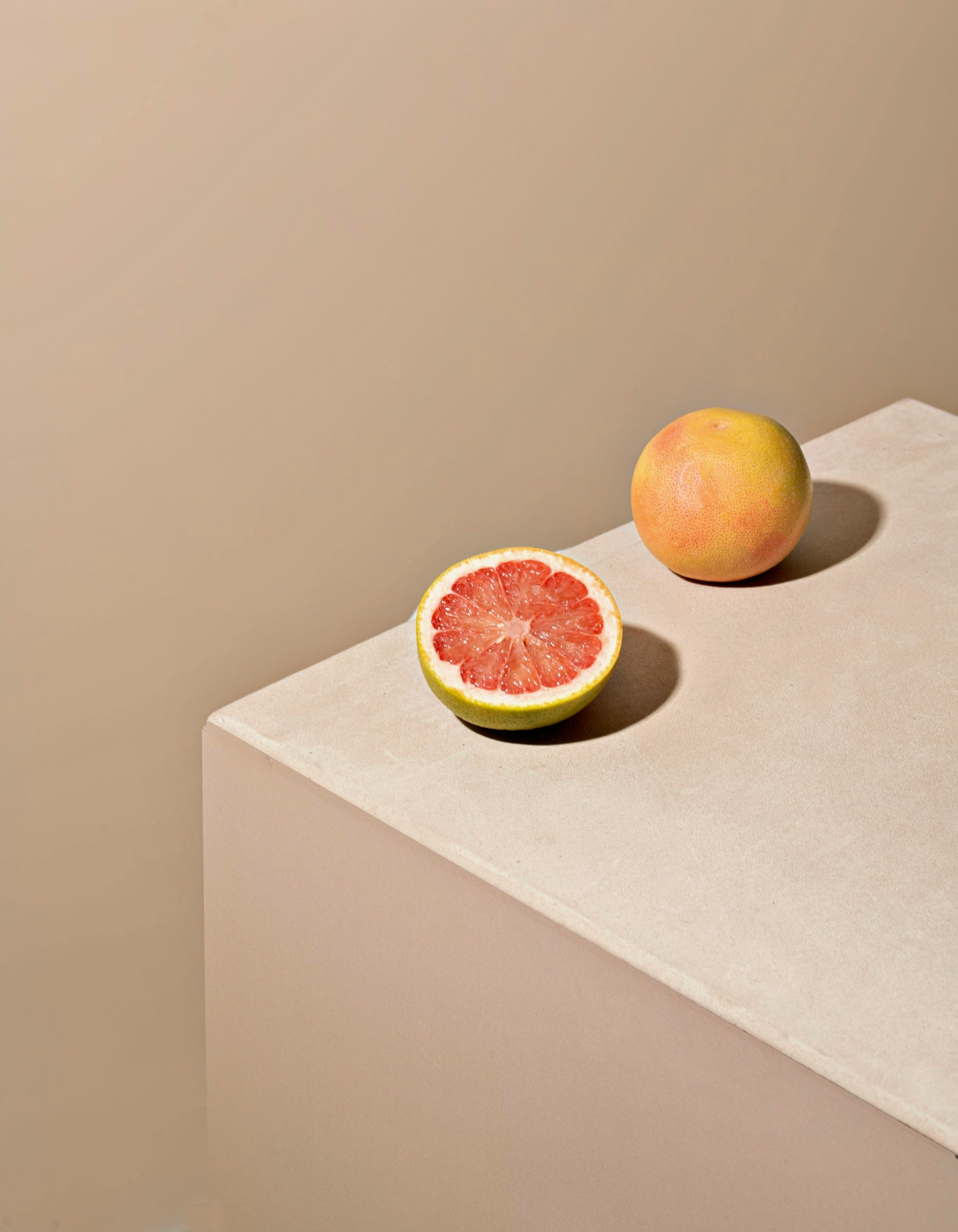 A whole grapefruit and a halved grapefruit showing the pink interior, placed on a white surface against a plain beige wall.
