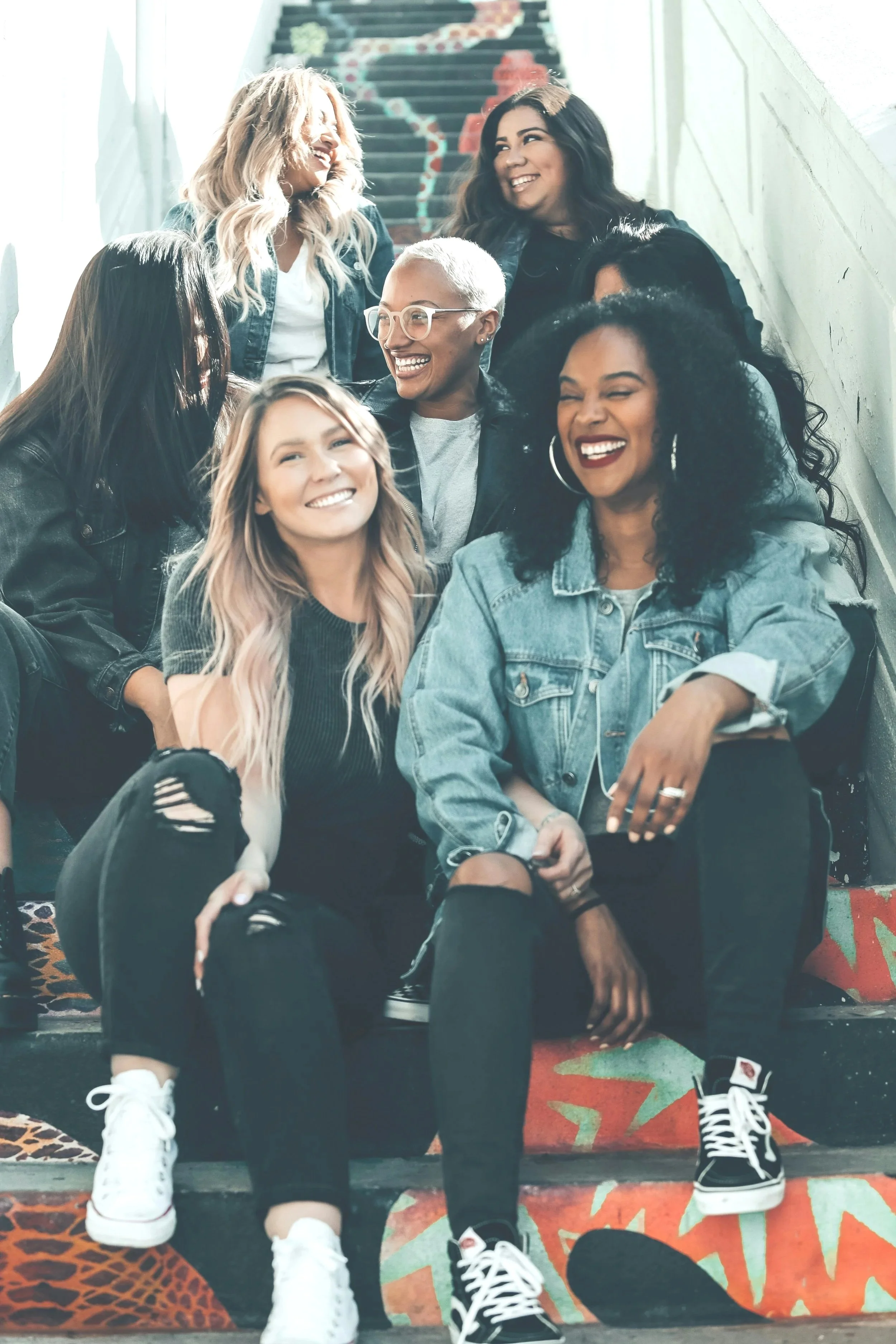 Group of women sitting on stairs, smiling and laughing together outdoors on a sunny day.
