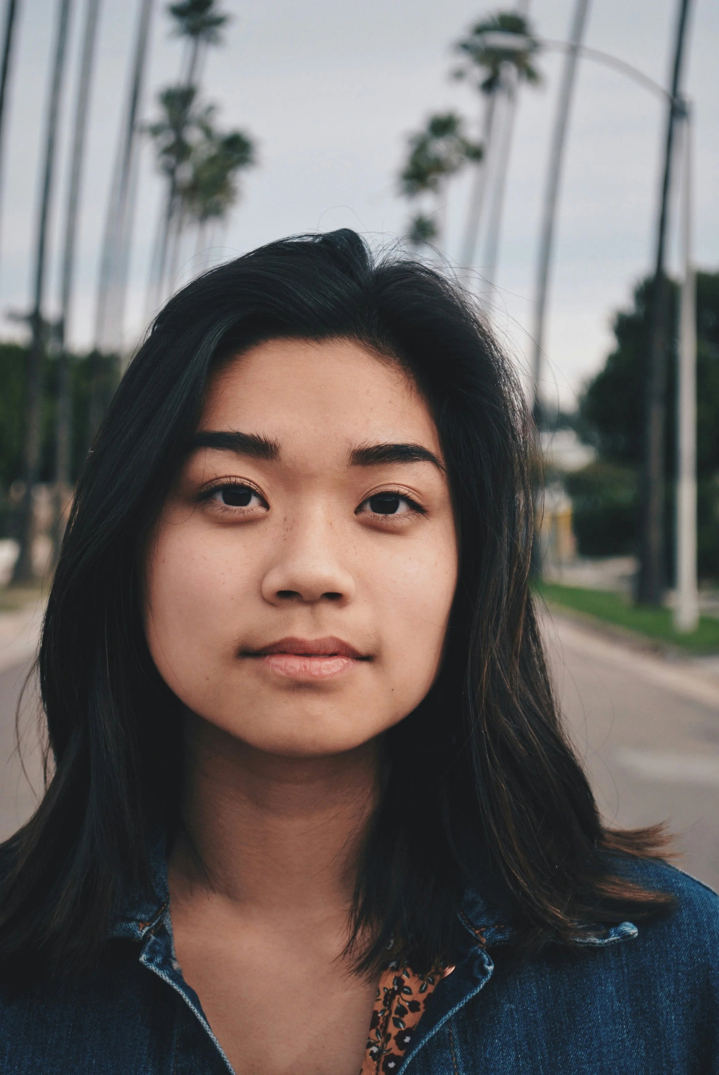 A young woman with black hair, brown eyes, and a light complexion. She is outdoors with tall palm trees and streetlights in the background, wearing a dark denim jacket over a patterned shirt.