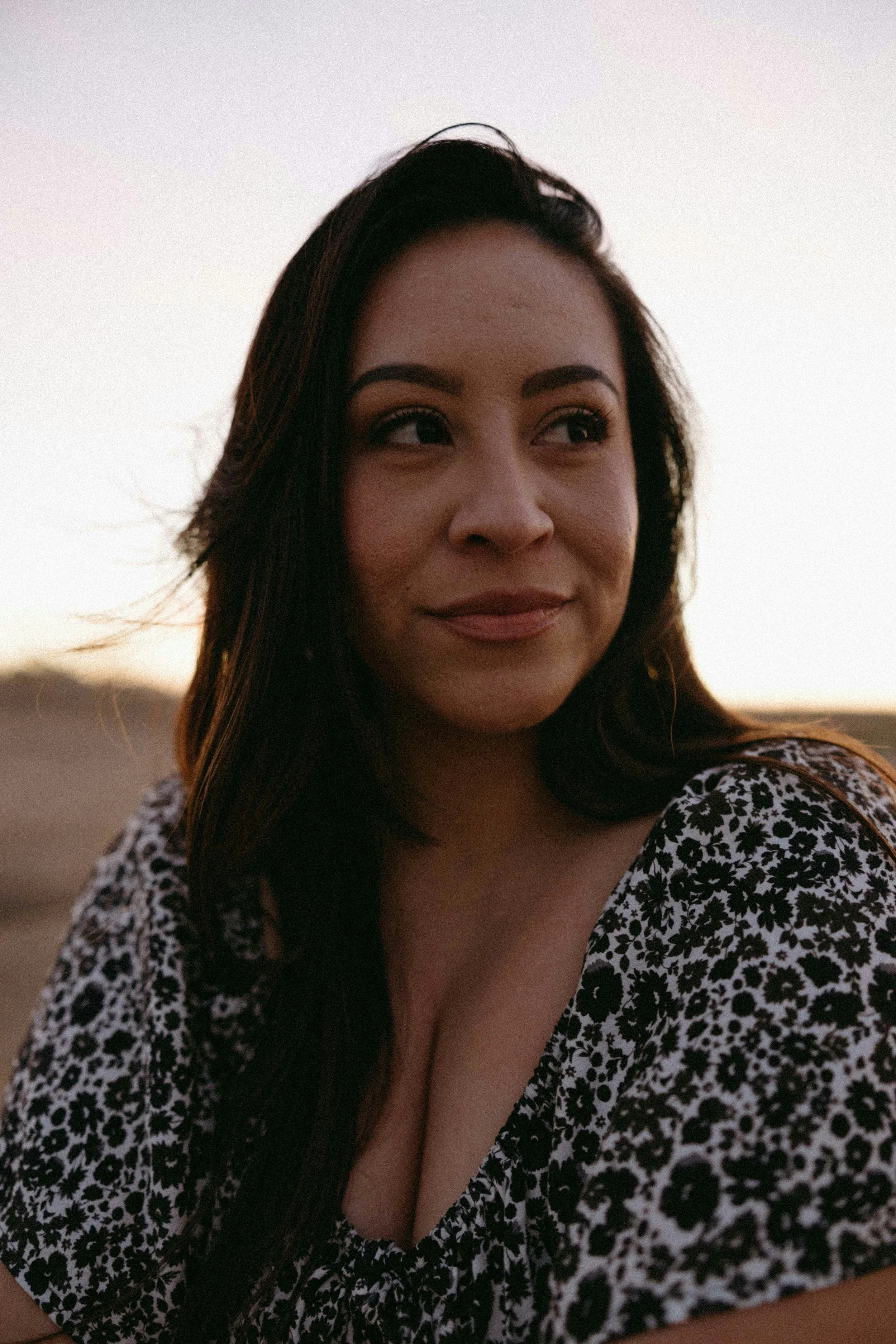 A woman with long dark hair, wearing a black and white floral dress, smiling outdoors during sunset.