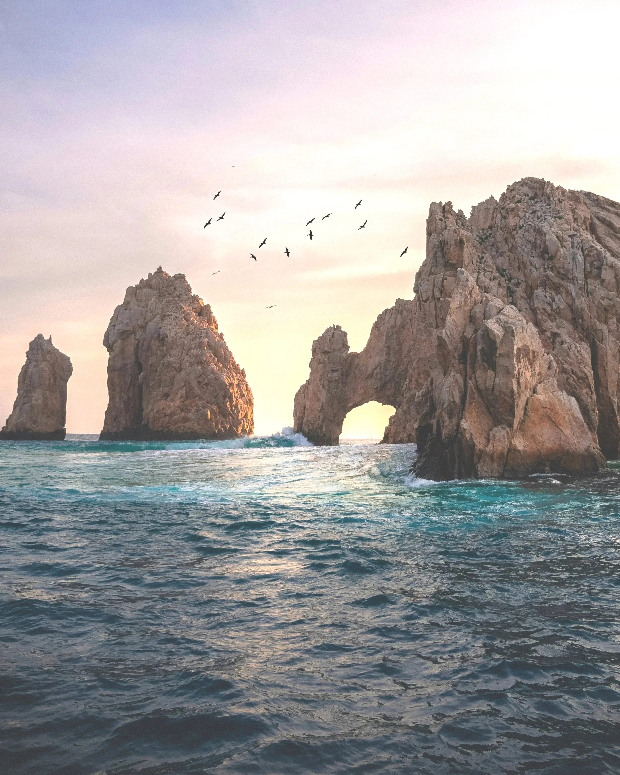 Ocean with rocky formations and a natural arch, birds flying in the sky during sunset.