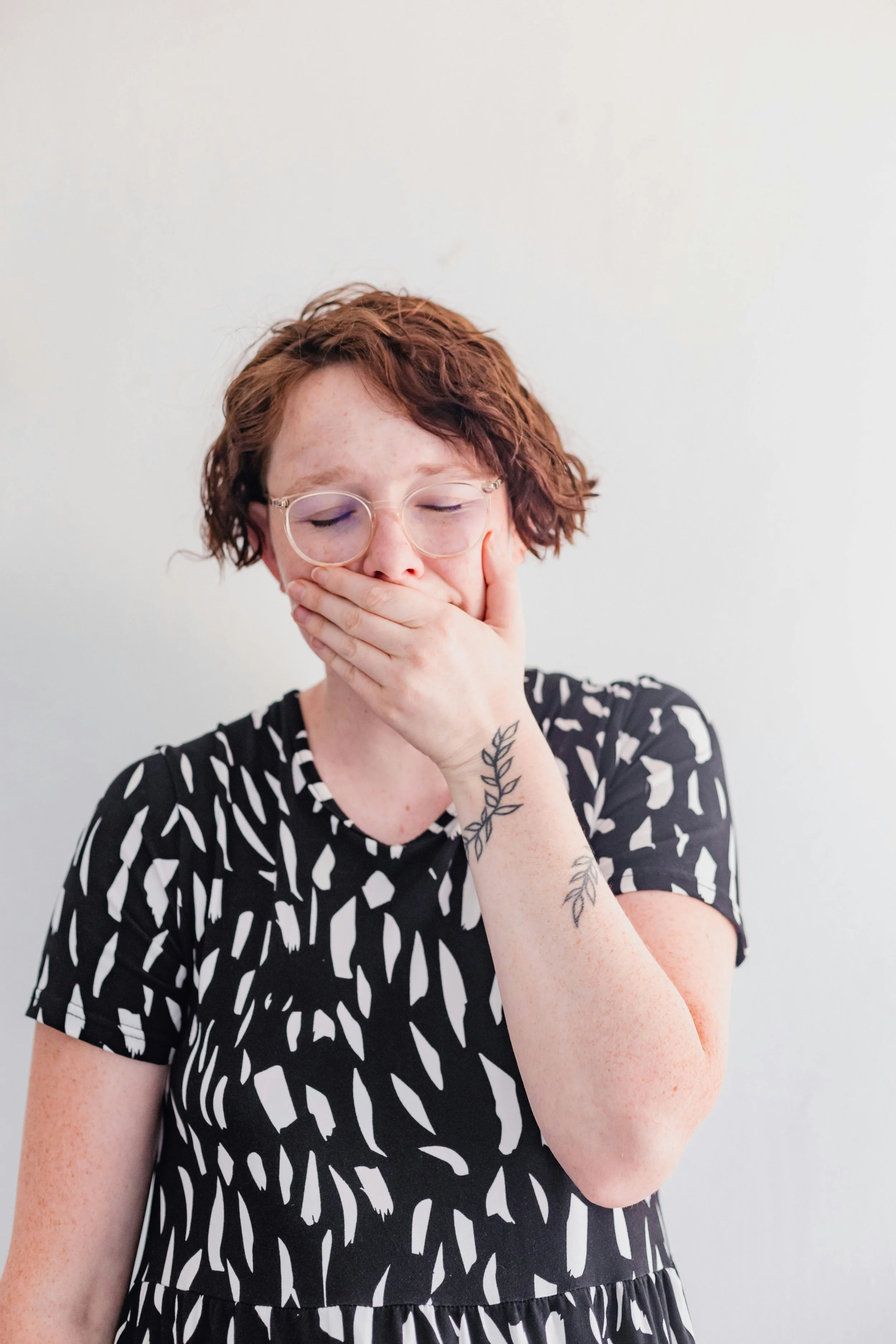 A woman with short curly red hair and glasses, wearing a black and white patterned dress, covers her mouth with her hand and appears to be yawning or surprised. She has a tattoo of leaves on her arm, and is standing against a plain white background.