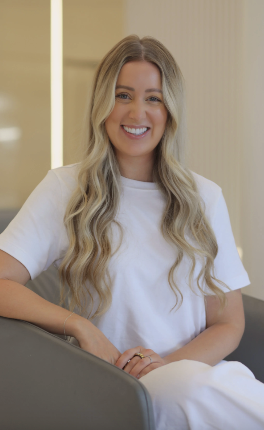 A smiling woman with long, wavy blonde hair wearing a white t-shirt sitting on a gray sofa in an indoor setting.