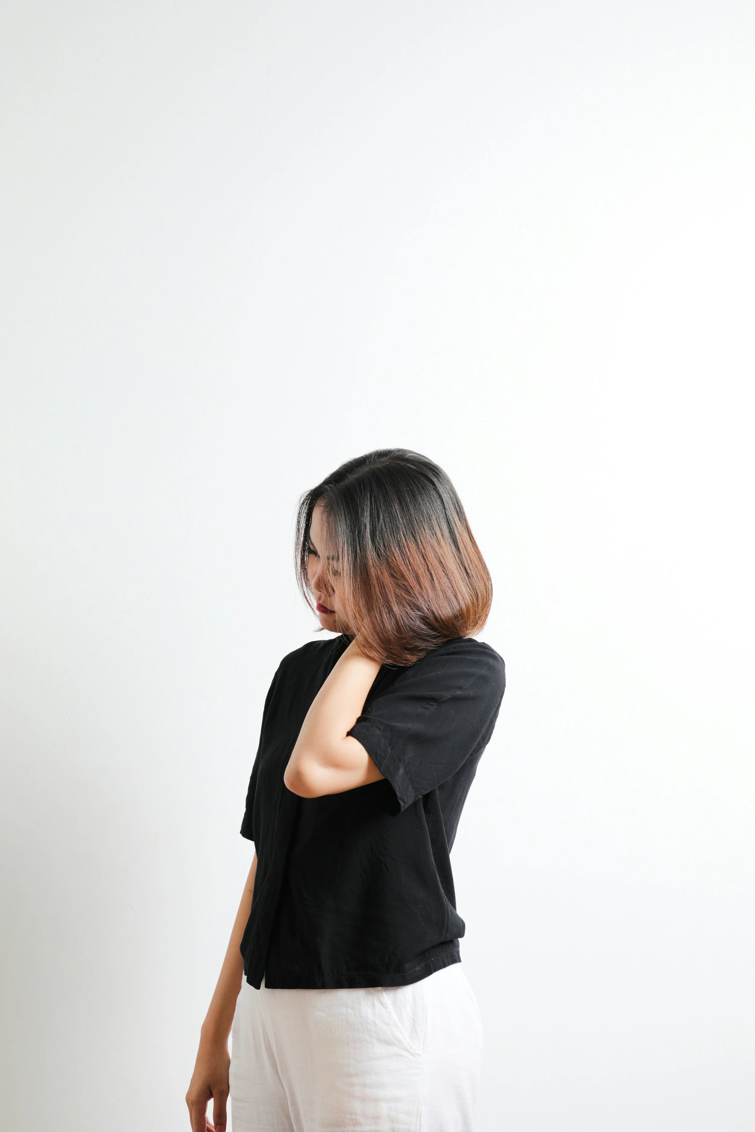 A woman with brownish black hair styled with a reddish tint at the ends, wearing a black top and white pants, standing against a plain white wall, with one hand touching her neck and her eyes closed.