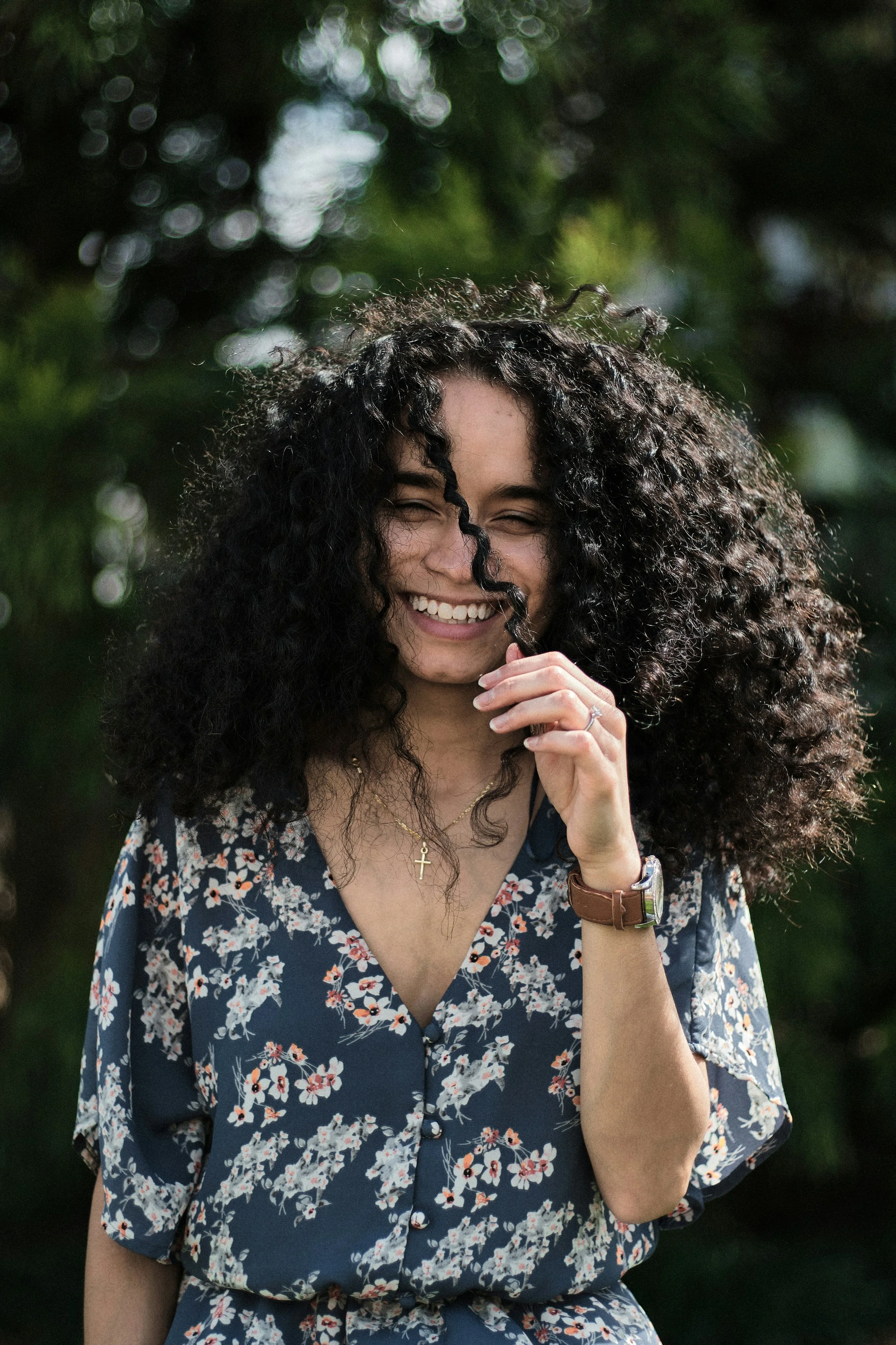 A woman with dark, curly hair smiling outdoors, wearing a floral dress, a cross necklace, watch, and bracelet, with a background of bokeh green foliage.