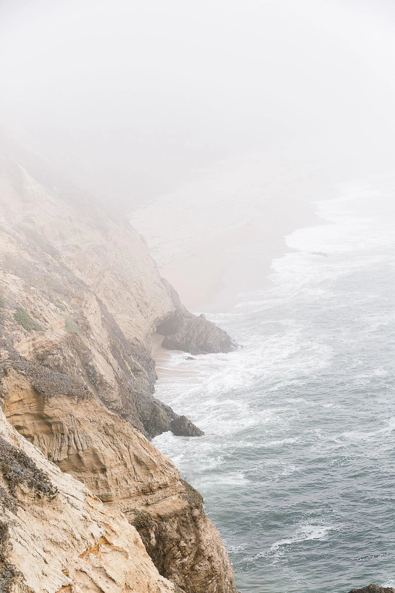 Cliffs along the coast with rocky terrain and ocean waves crashing at the base, overcast sky in the background.