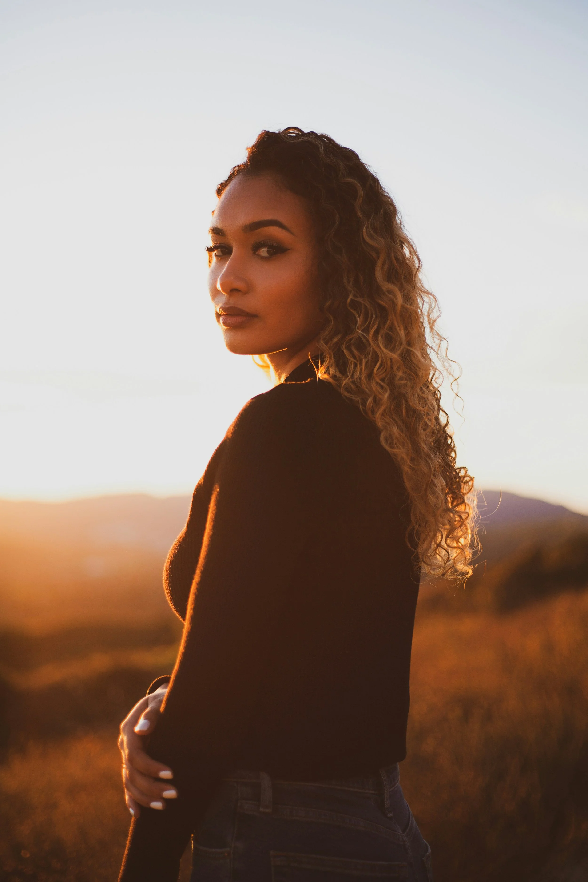 A woman with curly hair looking over her shoulder during sunset, standing outdoors with a landscape of mountains and a clear sky in the background.