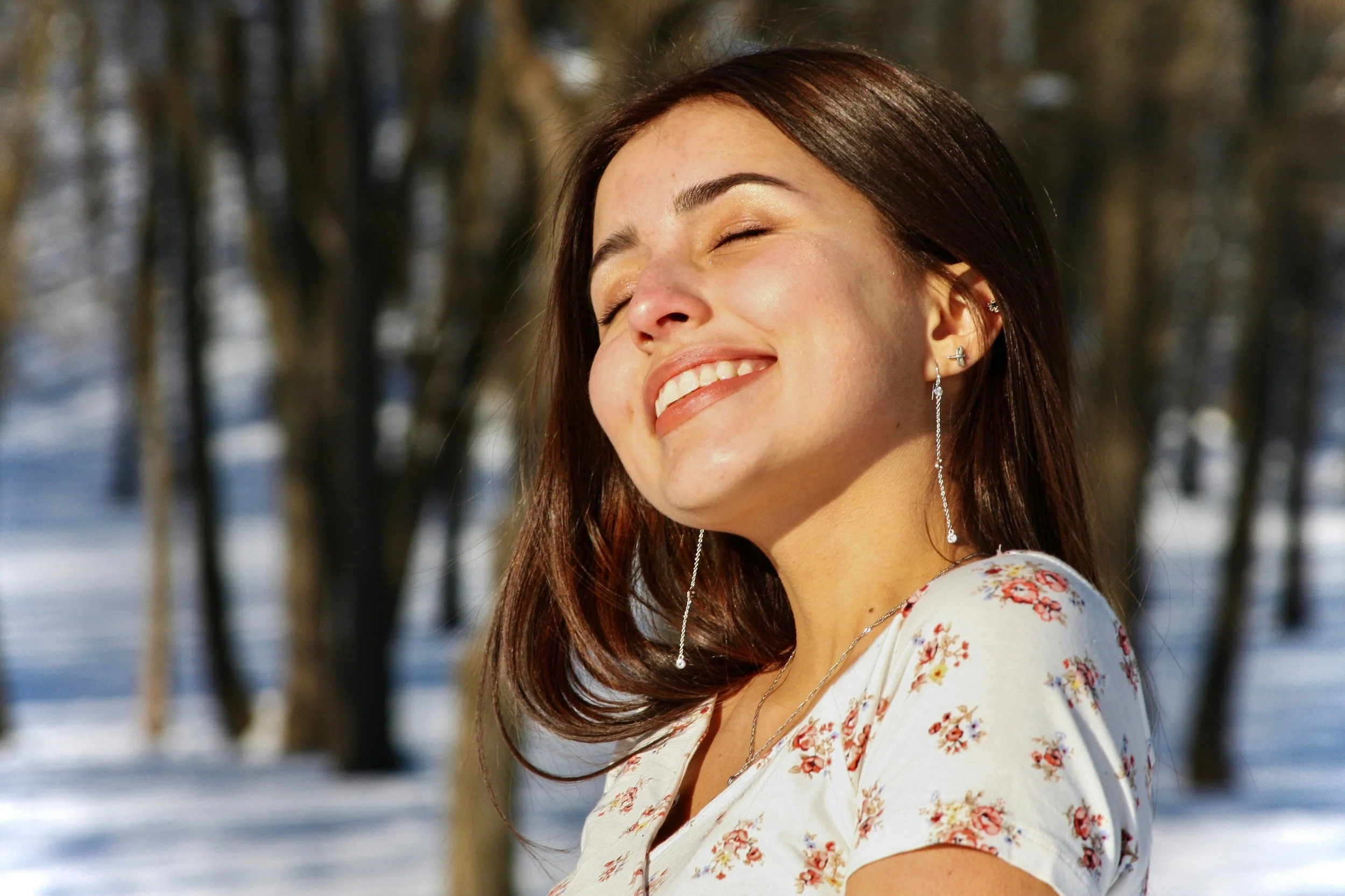 A woman with dark brown hair smiling and enjoying the sunlight outdoors in a snowy area with bare trees in the background.