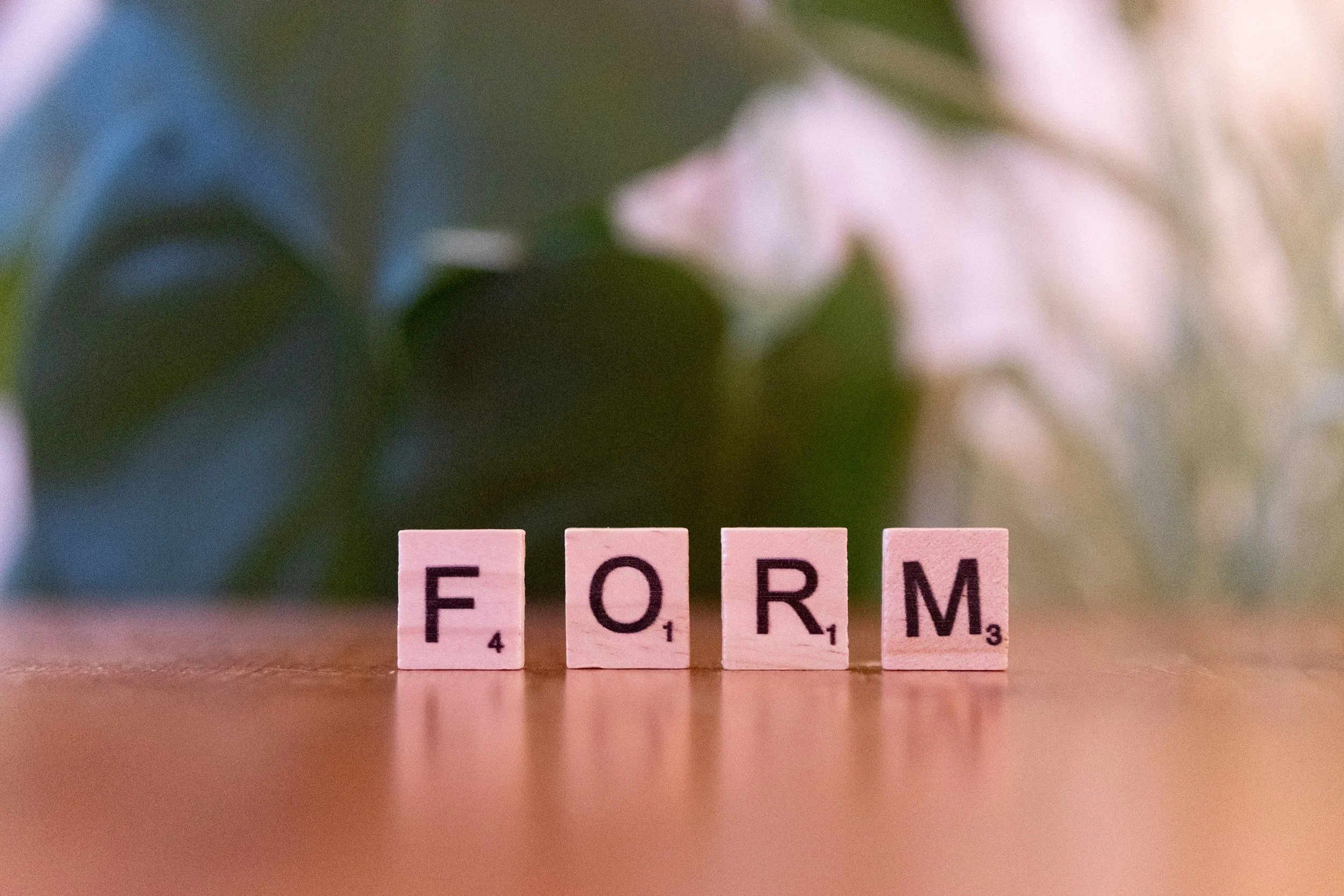 Close-up of pink Scrabble tiles spelling out the word 'FORM' on a wooden surface, with a blurred background of a flower.