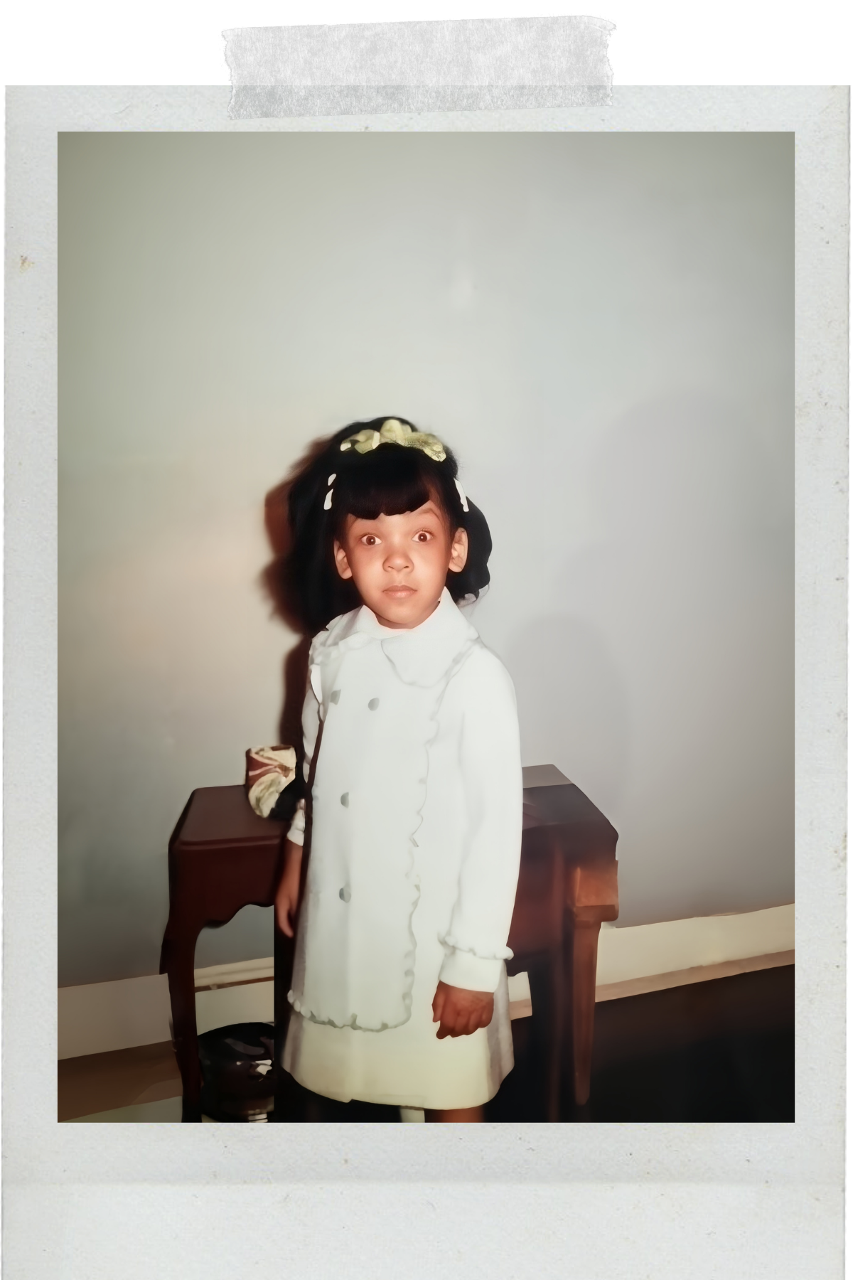 A young girl with dark hair and light skin, wearing a white coat with ruffles and large buttons, stands in front of a plain light-colored wall. She has a surprised or curious expression and a bow in her hair. A small wooden table is behind her.
