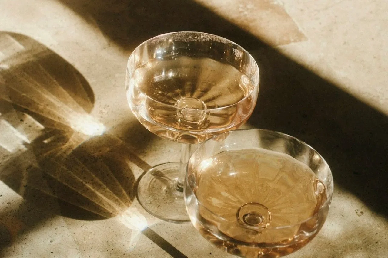 Two coupe glasses with pink liquid, placed on a textured surface, casting shadows and reflections.