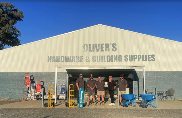 Group of five people standing in front of Oliver's Hardware & Building Supplies store with various equipment and tools outside.