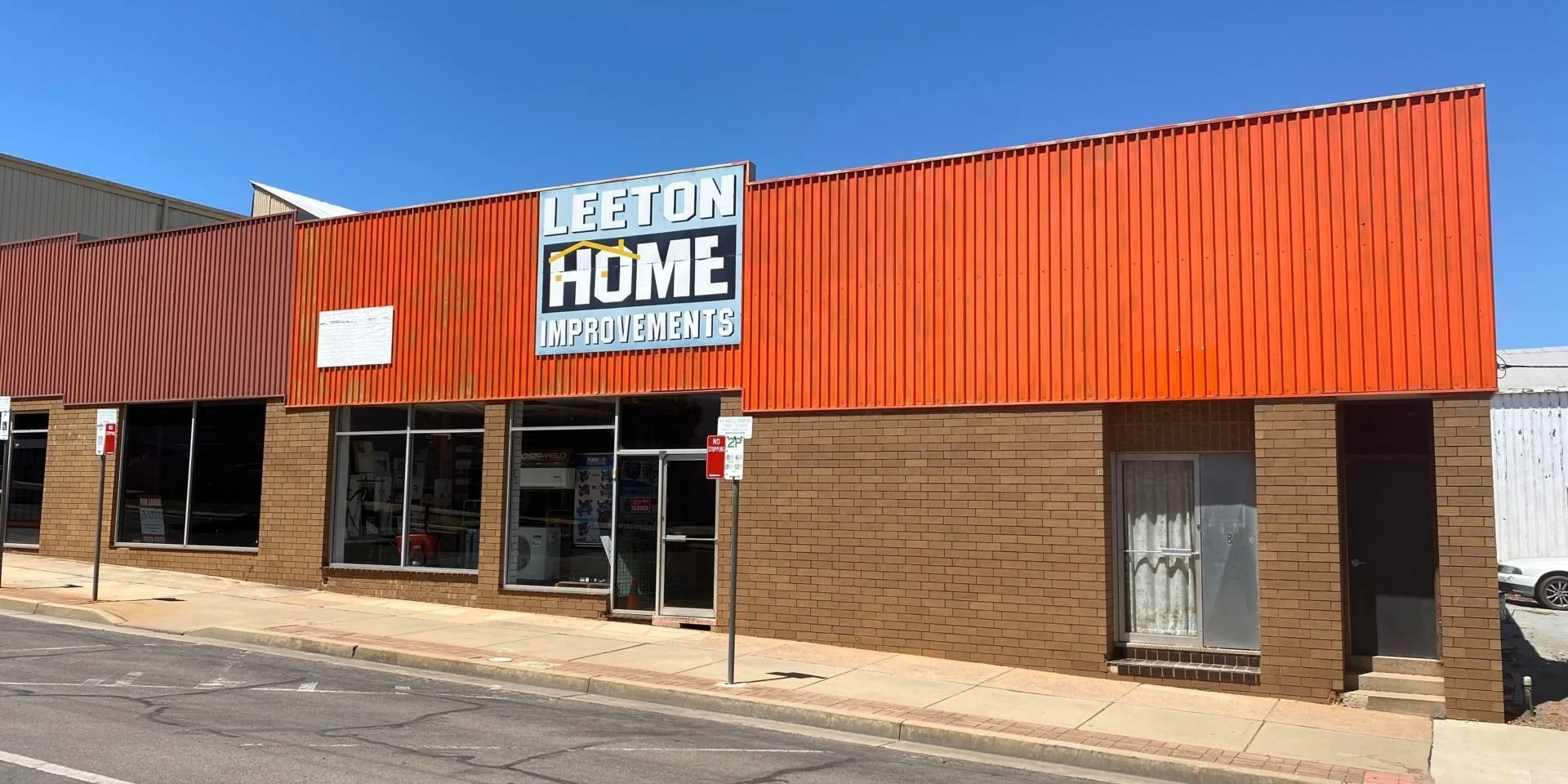 Storefront with orange siding and brick lower level, signage reads "Leeton Home Improvements" under a blue sky.