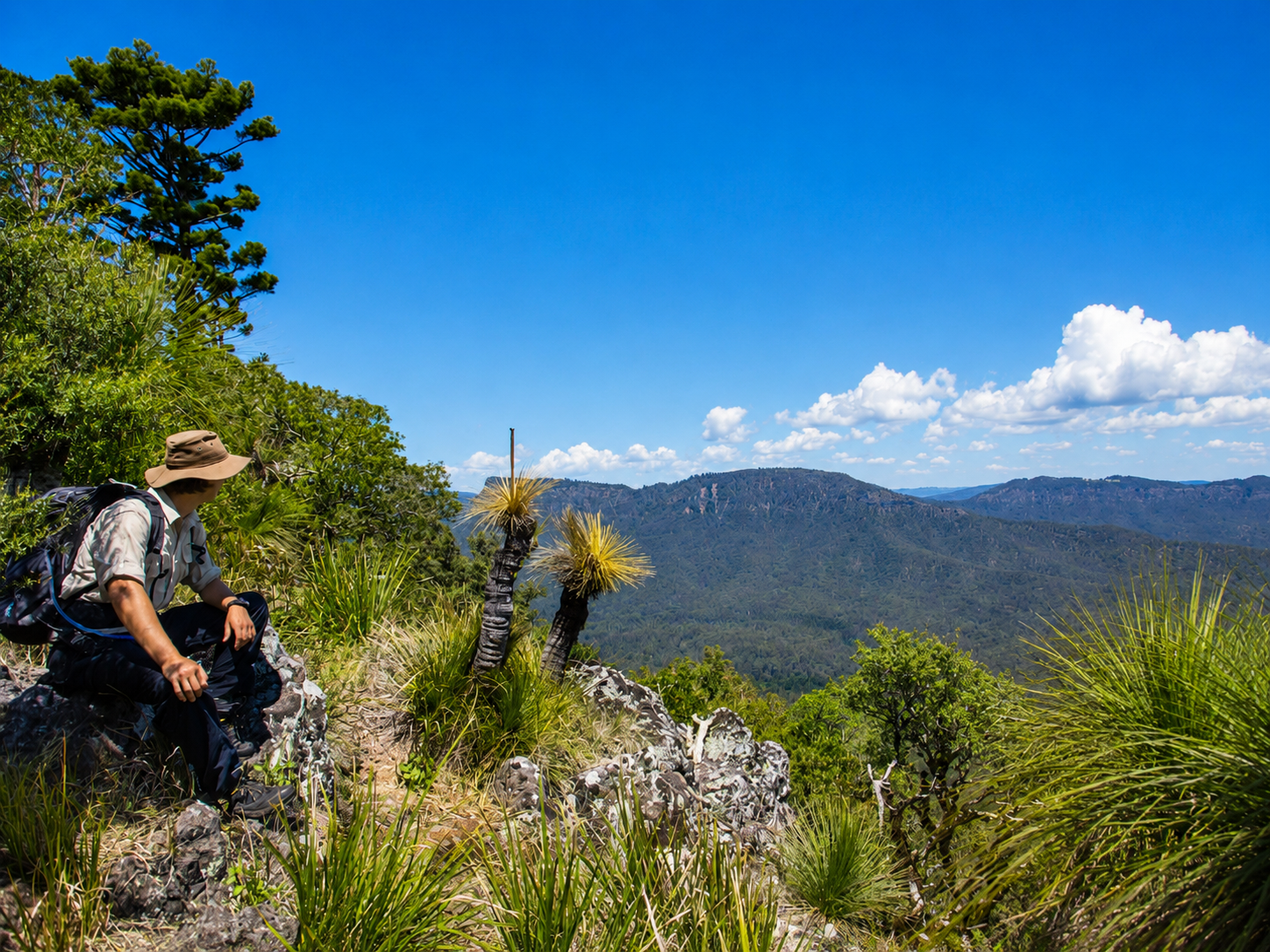 Hiker resting on rocky trail in lush green mountains under blue sky with white clouds.