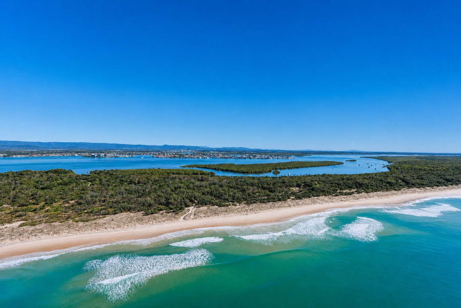 Aerial view of a beach with turquoise waves, sandy shoreline, lush green vegetation, and a large river or lake with boats in the distance under a clear blue sky.