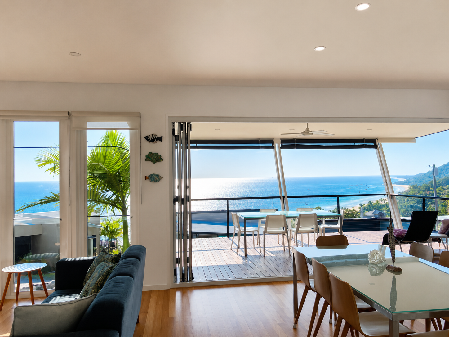 Living room with large windows and a view of the ocean and mountains, outdoor deck with dining table, chairs, and glass walls, modern decor with a black sofa, dining chairs, and tropical plants.
