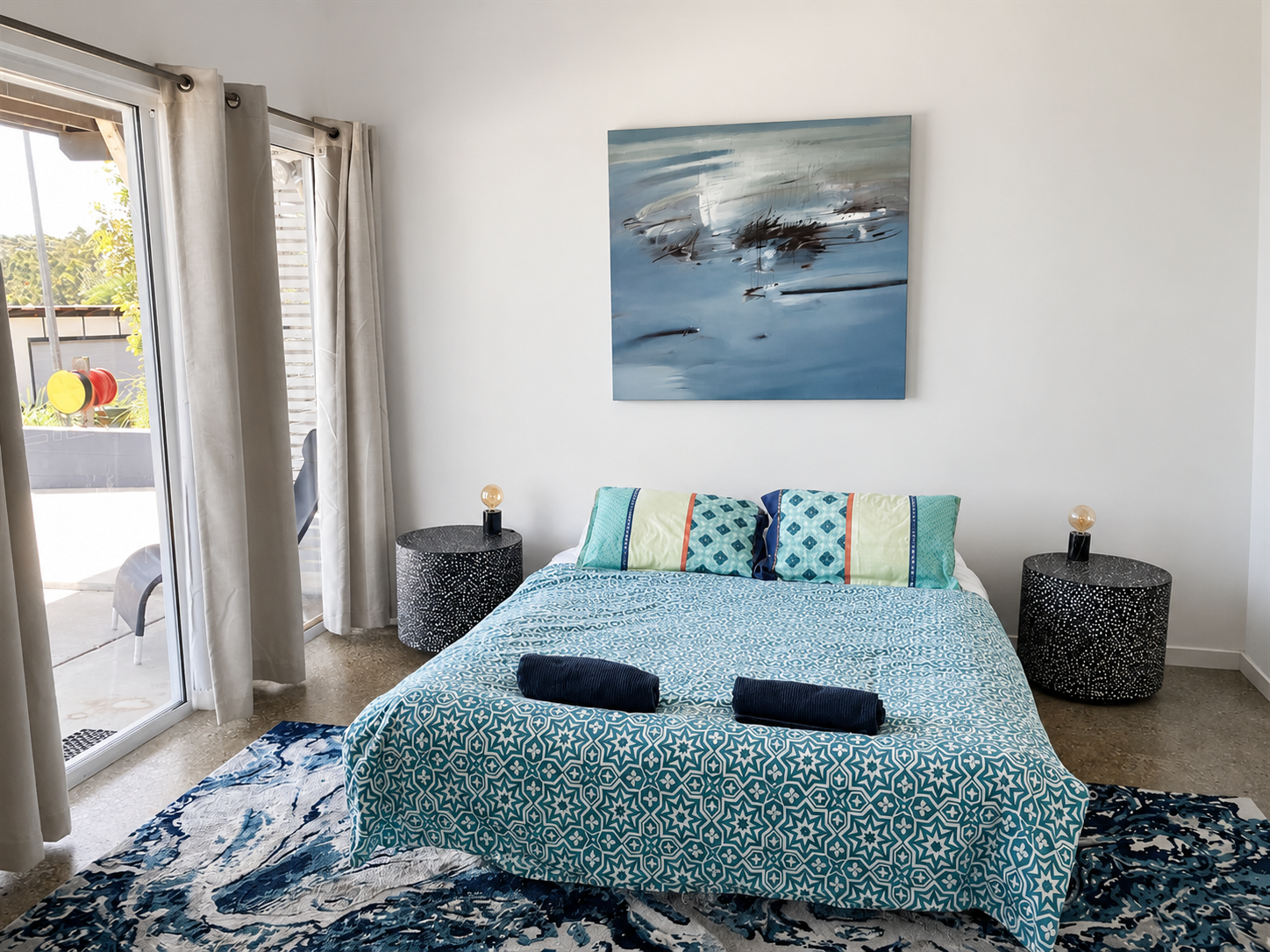 A bedroom with a bed covered in blue and white patterned bedding, two matching patterned pillows, and two folded dark blue towels on top. There are two black and white bedside tables with small lamps, a large abstract blue painting on the white wall 