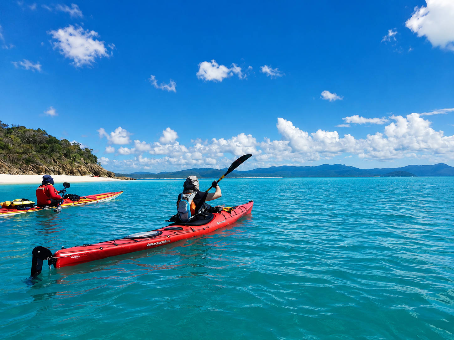 Two people kayaking on a calm blue ocean under a partly cloudy sky, with a green rocky coastline to the left and distant mountains in the background.