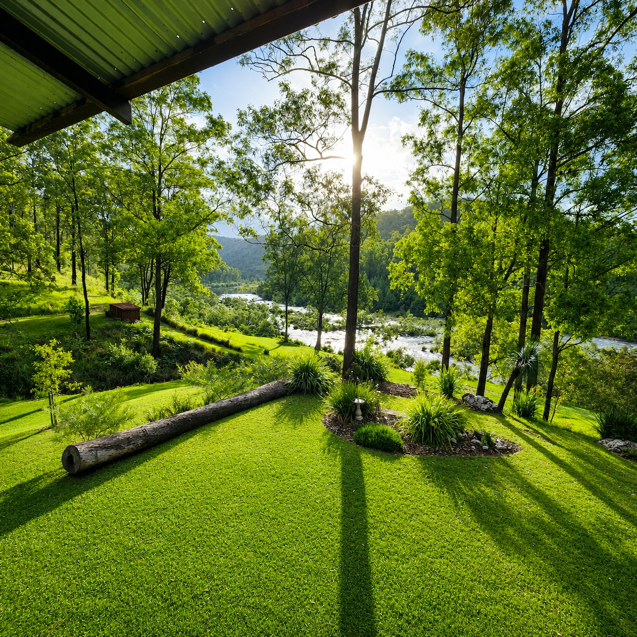 Sunlit backyard with lush green grass, trees, and landscaped plants overlooking a river, with hills in the background.