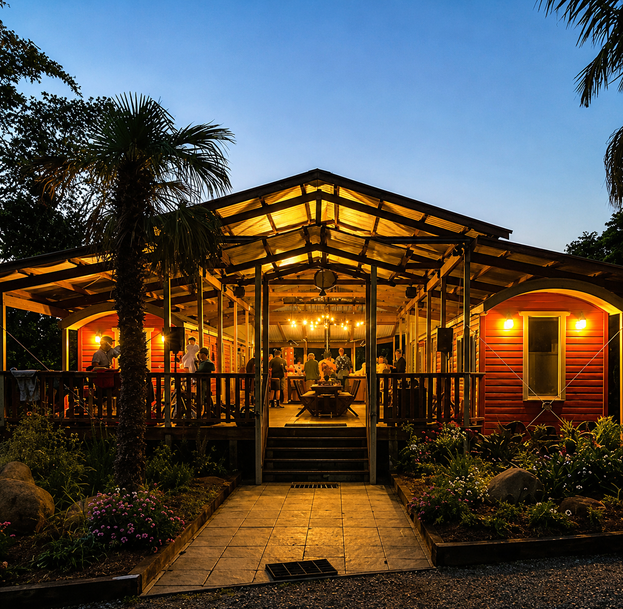 A lively outdoor gathering during the evening on a wooden deck attached to a red house, lit by warm string lights and wall sconces, with people socializing and a garden with flowers in the foreground.
