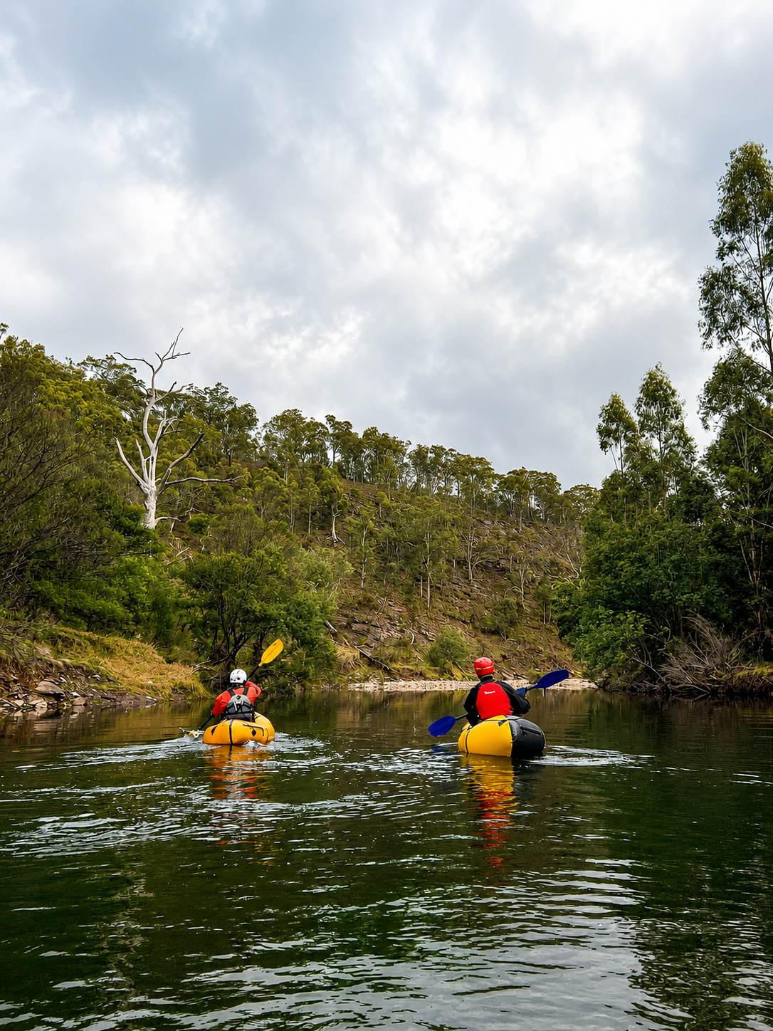 Two people in yellow kayaks paddling on a river with forested hills and cloudy sky in the background.