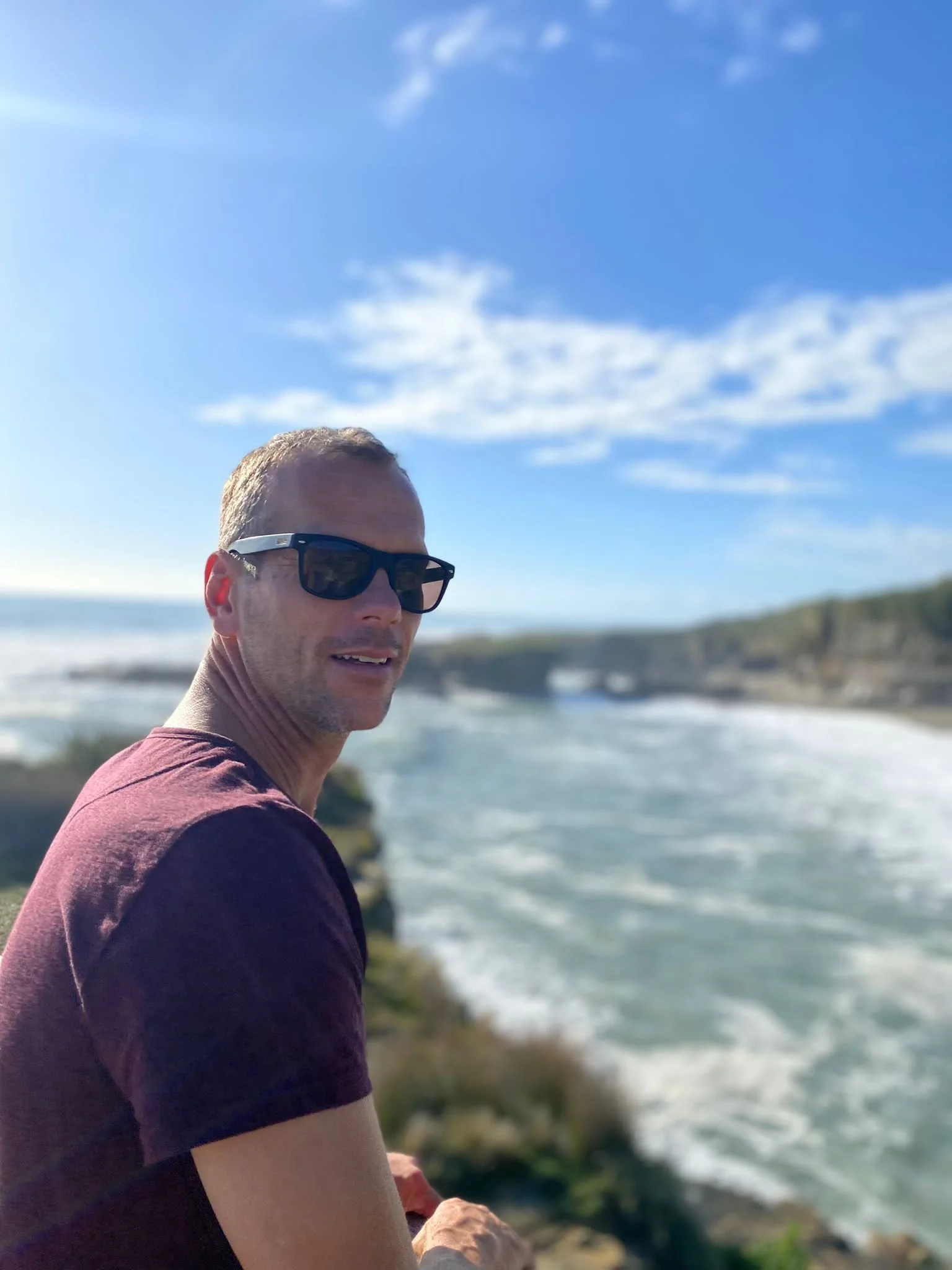 A man wearing sunglasses sitting near the ocean on a sunny day, with blue sky and white clouds overhead, and cliffs in the background.