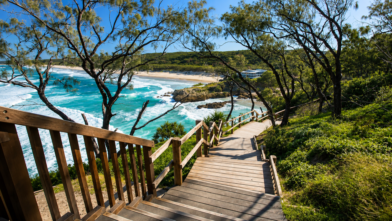 Wooden stairs and railing leading down a hillside toward a sandy beach with rocky outcrops and turquoise ocean waves, surrounded by green trees and foliage under a clear blue sky.