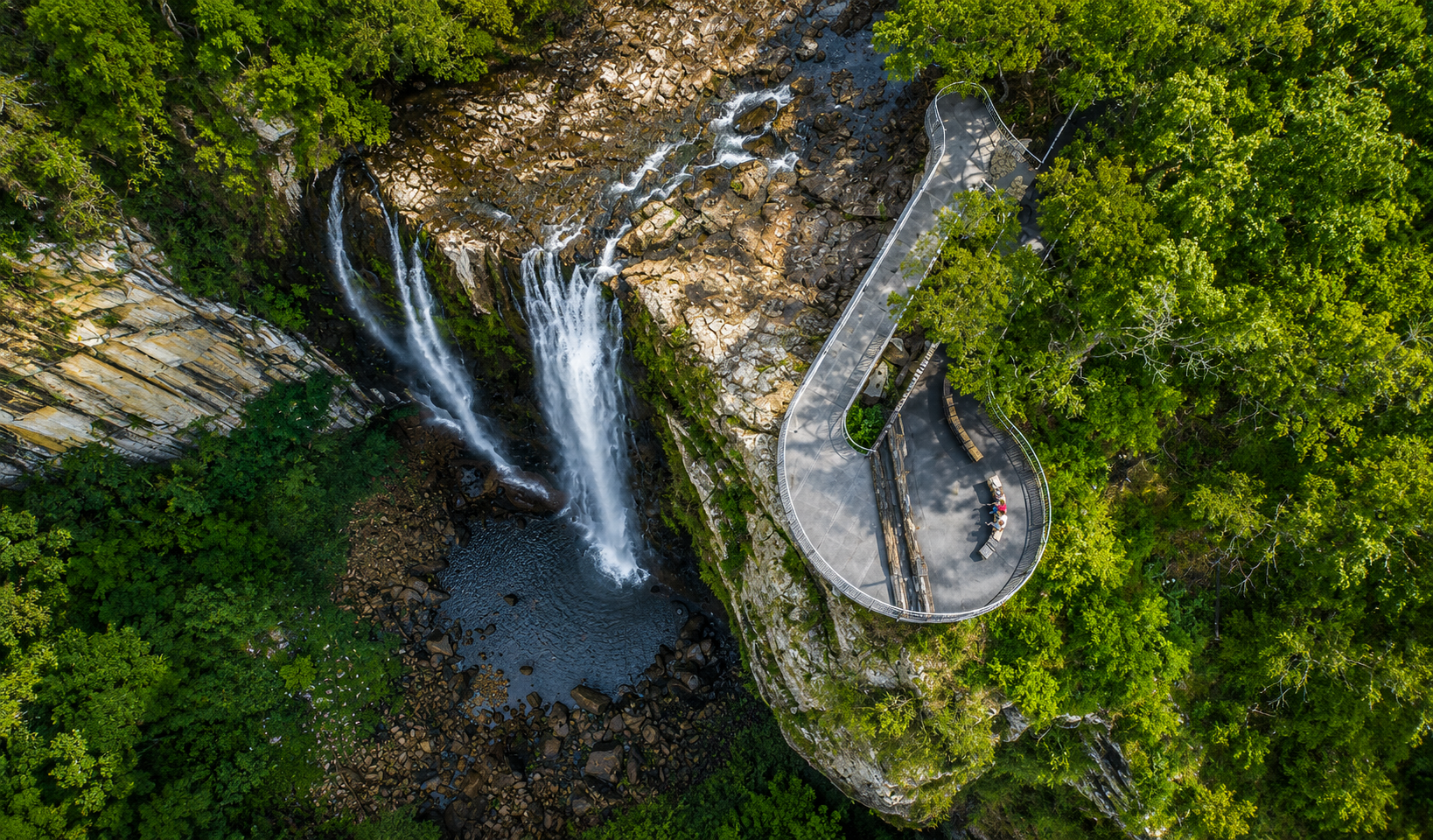 Aerial view of a waterfall flowing down a rocky cliff surrounded by green trees, with a curved viewing platform and benches on a walkway overlooking the waterfall.