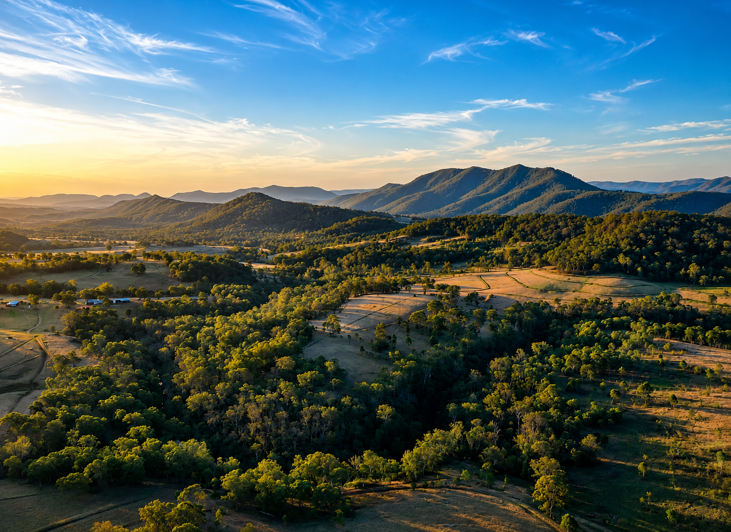 A scenic aerial view of rolling green hills and mountains at sunrise with scattered trees, fields, and a few farm buildings