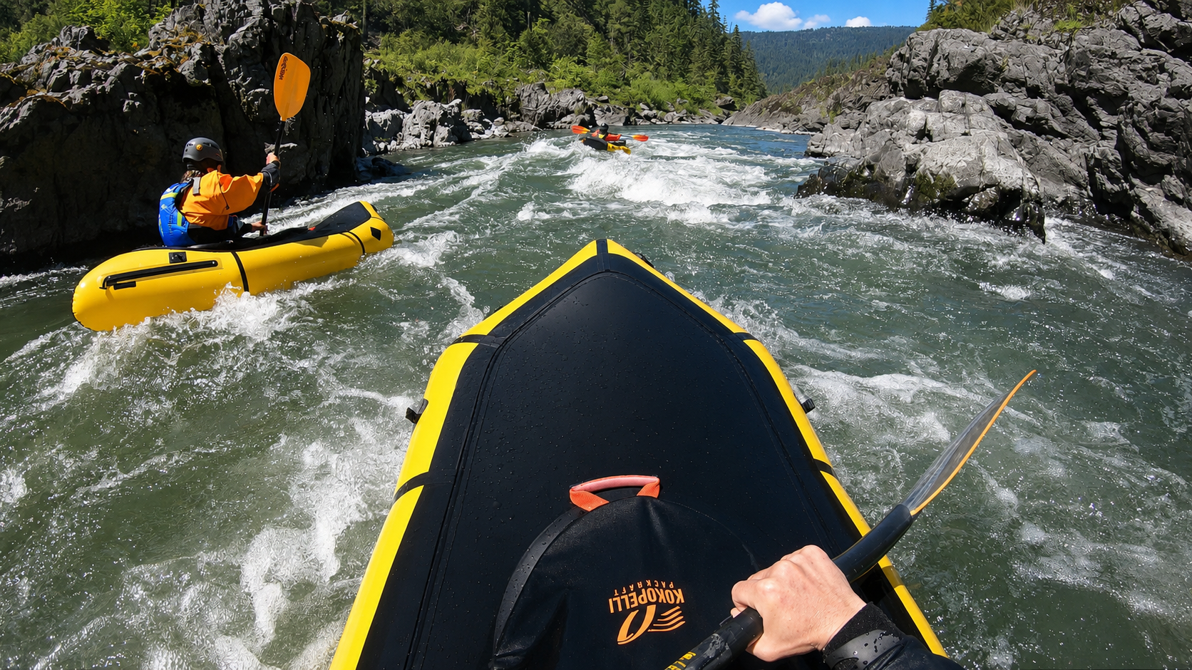 Two people kayaking in a river surrounded by rocks and green trees, with mountain hills in the background on a sunny day.
