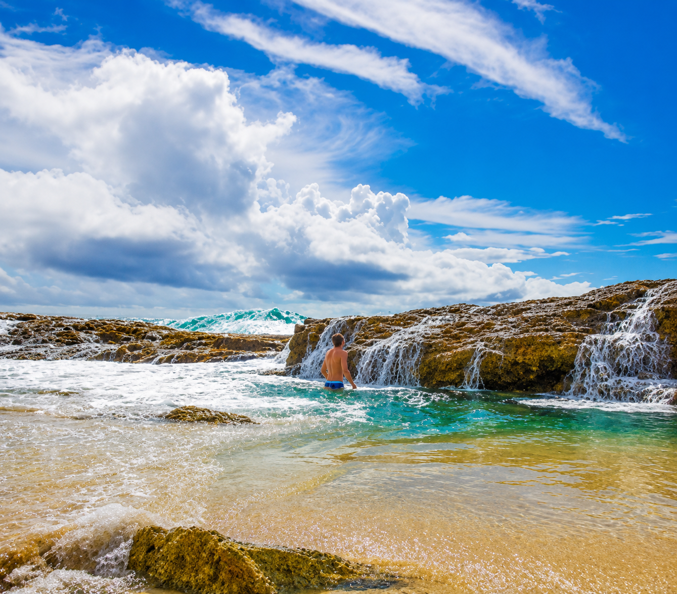 Man standing in the ocean near rocks on a sunny day with clouds and sky in the background.