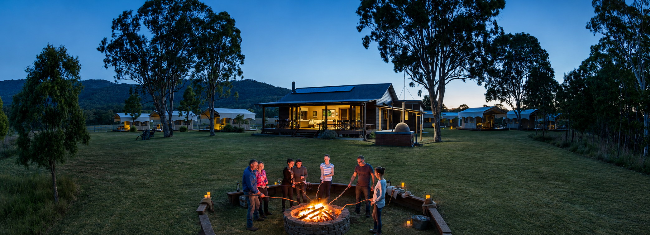 People gather around a campfire in a grassy open area during twilight, with a house and several tents or cabins in the background, surrounded by trees and hills.