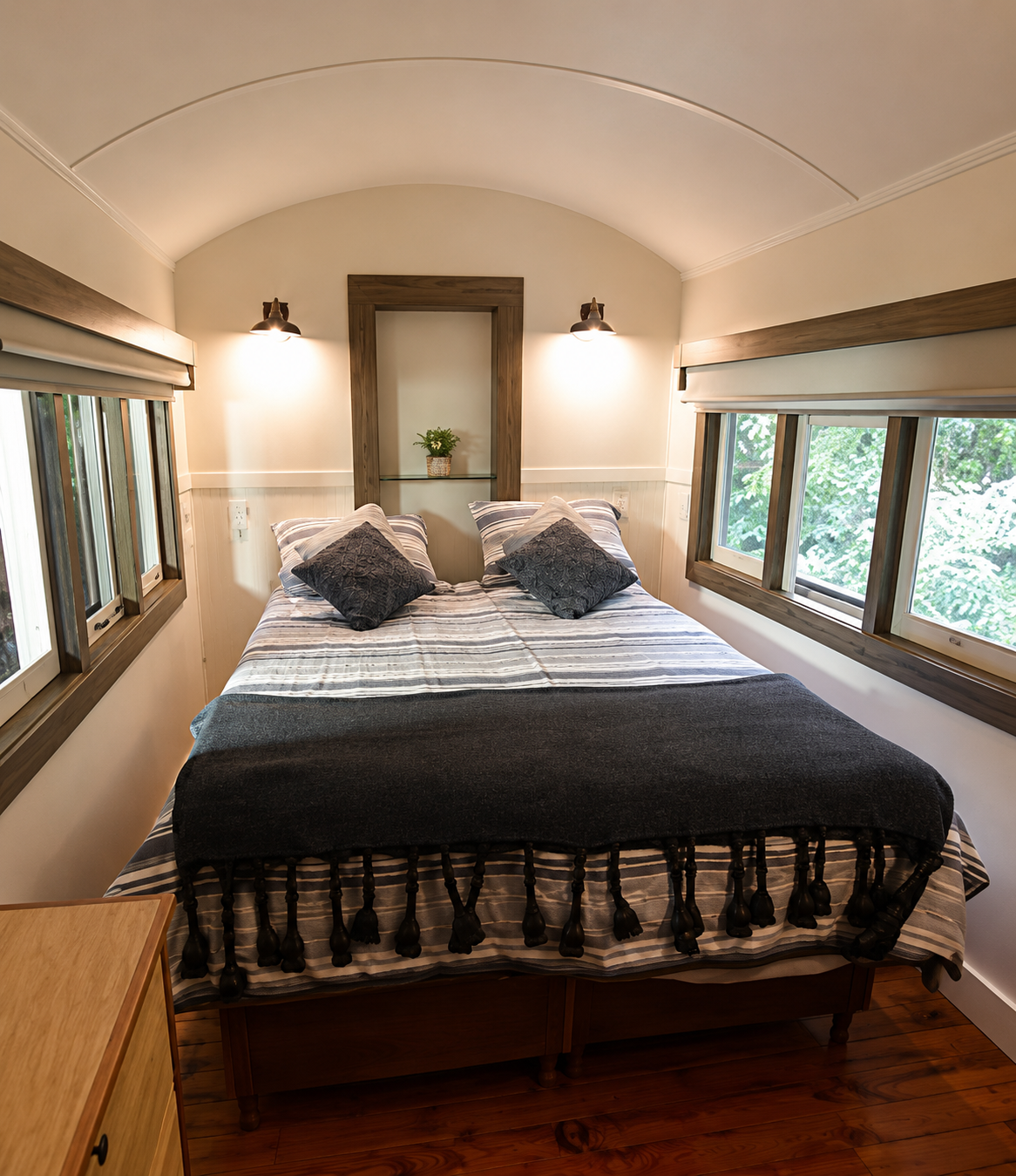 A cozy bedroom with a bed, black pillows, striped bedding, and wooden window frames, with greenery visible outside the windows.
