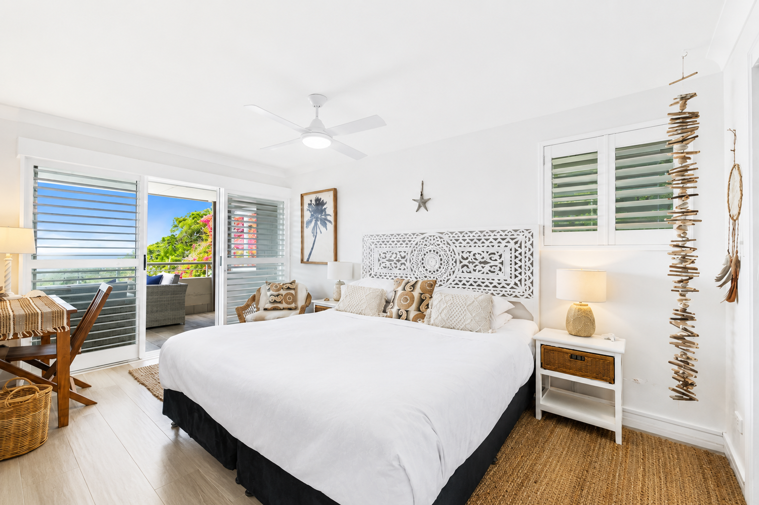 Bright bedroom with white walls, large bed with white bedding and decorative pillows, nightstands with lamps, wall art, and a ceiling fan. Balcony with outdoor seating visible through open sliding doors, decorated with beach-themed and natural decor 