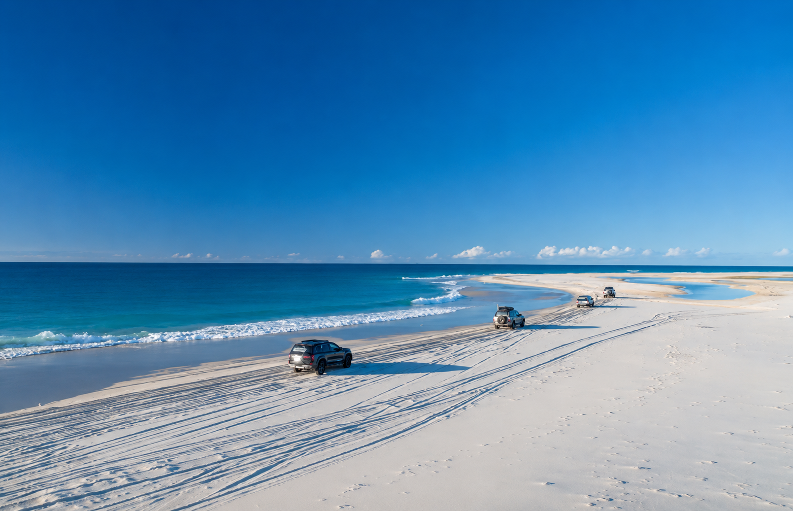 Four vehicles parked on a sandy beach with tire tracks, facing the ocean under a clear blue sky, with waves gently crashing on the shore.