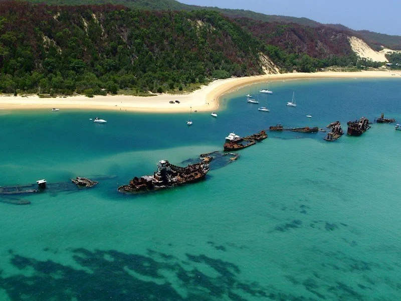 Aerial view of a wrecked ship near a sandy beach with green islands and blue waters, some boats anchored nearby.