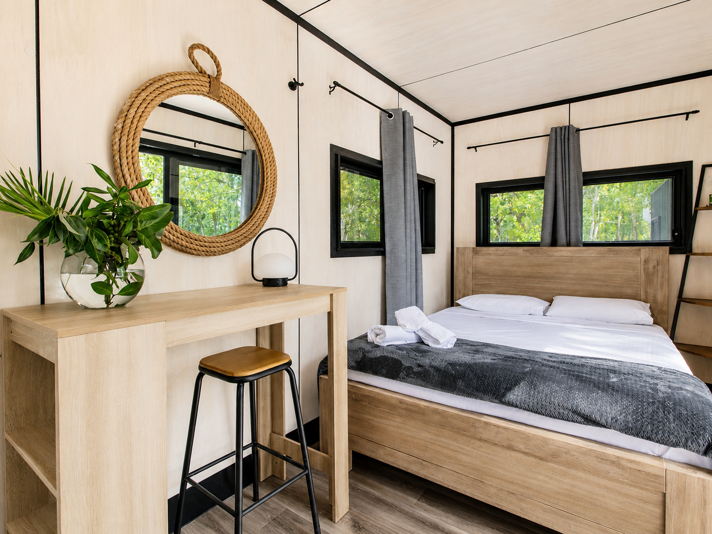 A cozy bedroom with a wooden bed, gray curtains, and three windows showing green trees outside. A wooden desk with a chair, a mirror with a rope border, a vase with green leaves, and a small lamp are next to the bed.