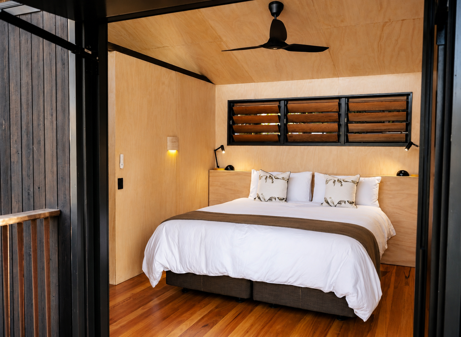 A modern bedroom with a wooden interior, featuring a bed with white bedding and decorative pillows, black wall-mounted reading lamps, a ceiling fan, and a black horizontal window with wooden slats.