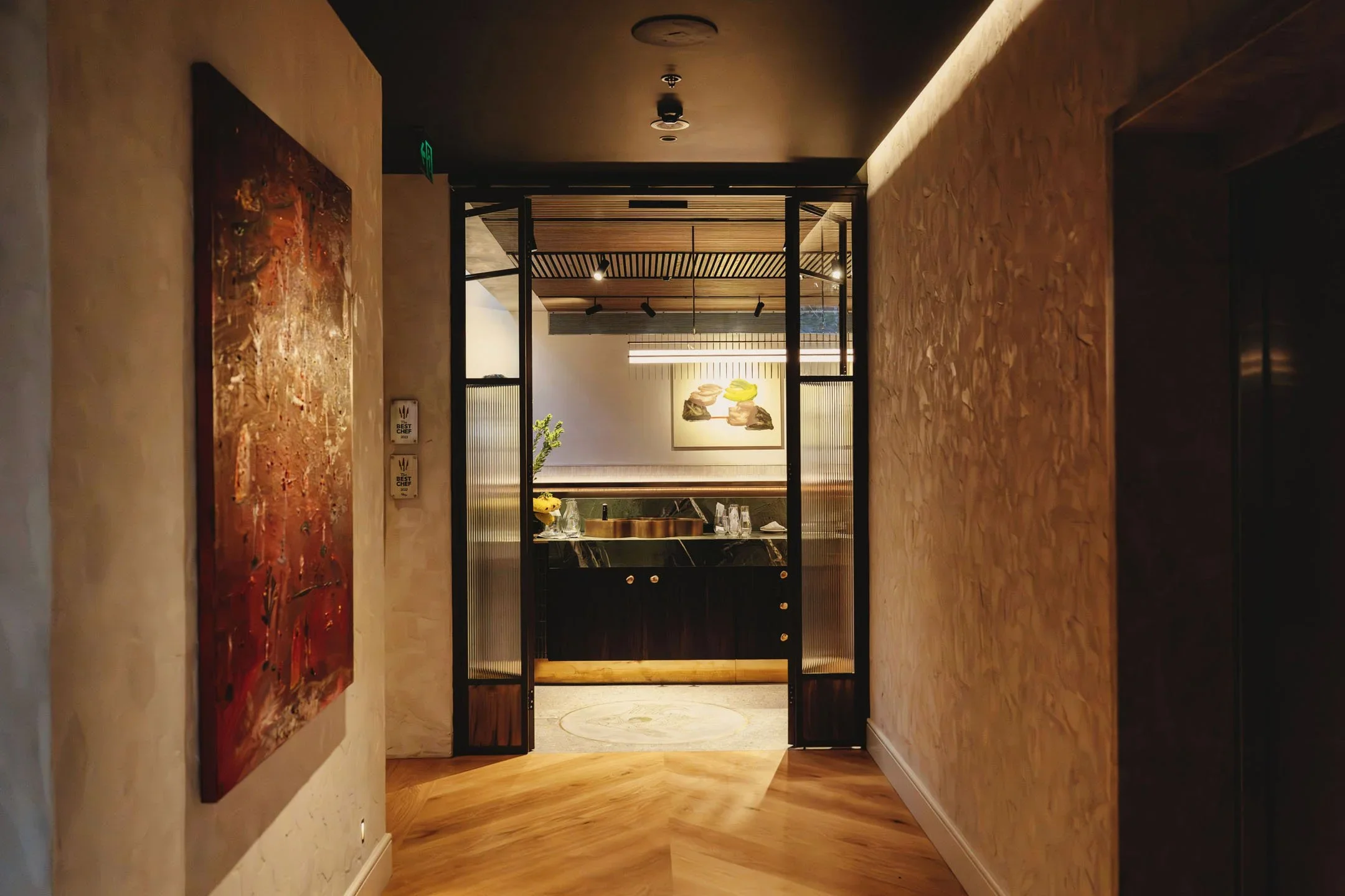 Interior view of a modern room with warm wooden floors, beige textured walls, and a large abstract painting on the left wall. In the background, there is a dining or lounge area with a decorative sculpture, greenery, and lighting fixtures.